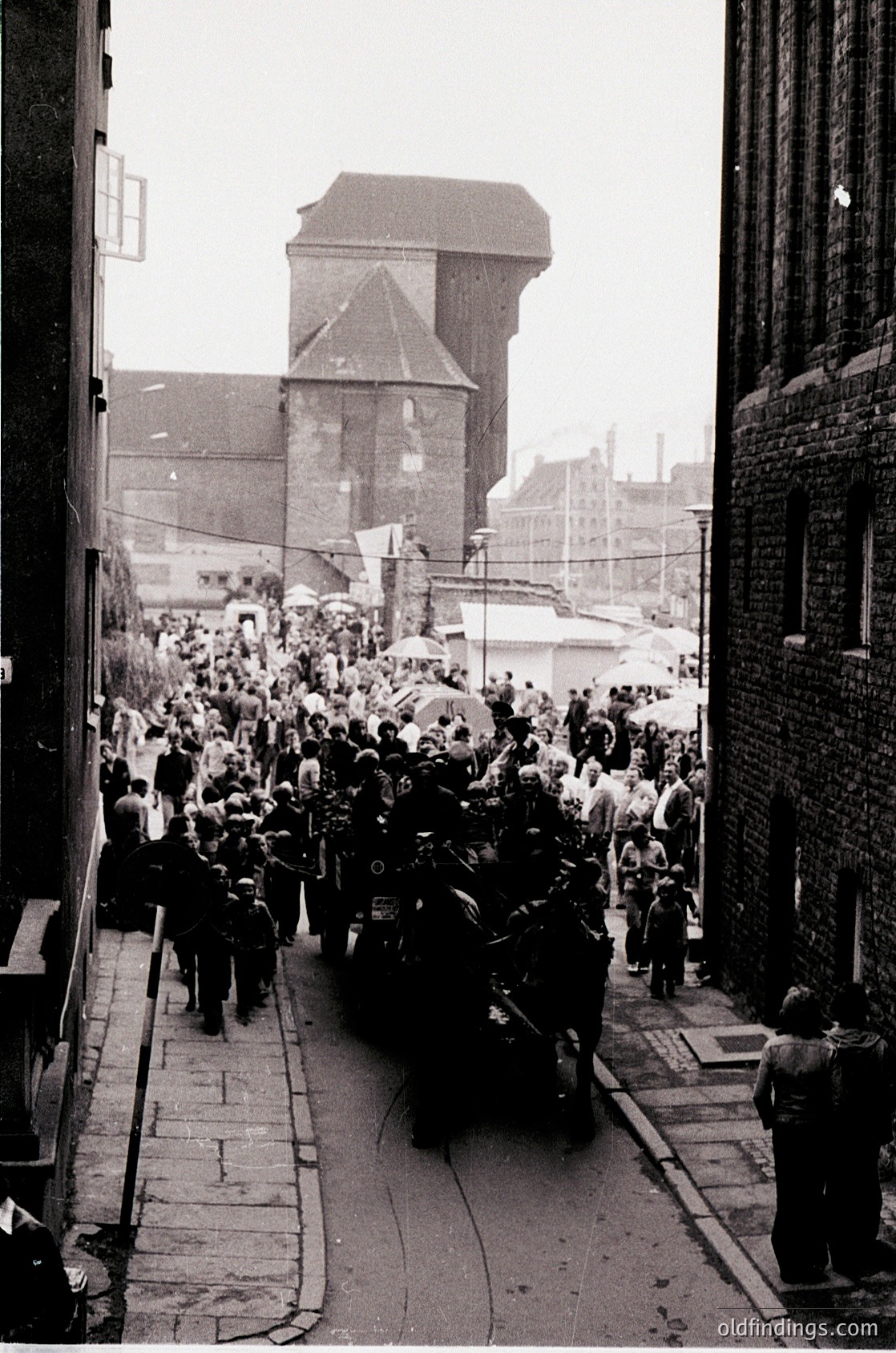 Crowded urban procession in a narrow alleyway, flanked by brick buildings and industrial architecture. A hearse carries a flag-draped coffin, followed by uniformed personnel and mourners. Prominent brick tower in background suggests historical European cityscape. Likely mid-20th century funeral procession.