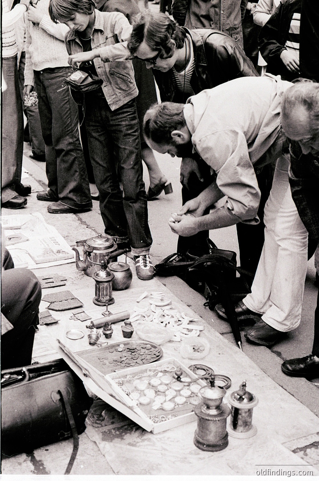 Street-side vintage repair stand with brass and copper tools, featuring a cobbler mending boots while surrounded by antique shoe polish pots and vintage footwear. Crowd in 1960s-70s urban setting, likely Europe.