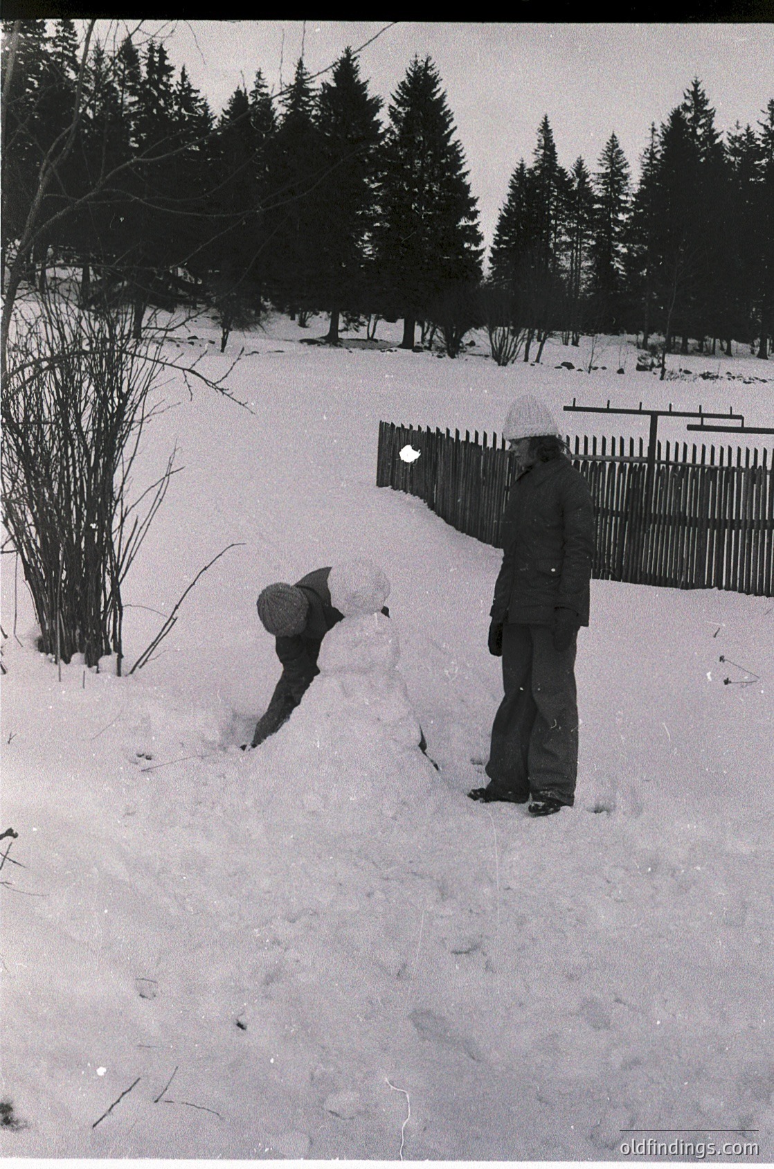Black-and-white snapshot of two children building a snowman in a residential backyard, surrounded by snow-covered trees and a wooden fence. Clothing suggests mid-20th century winter attire ( ).