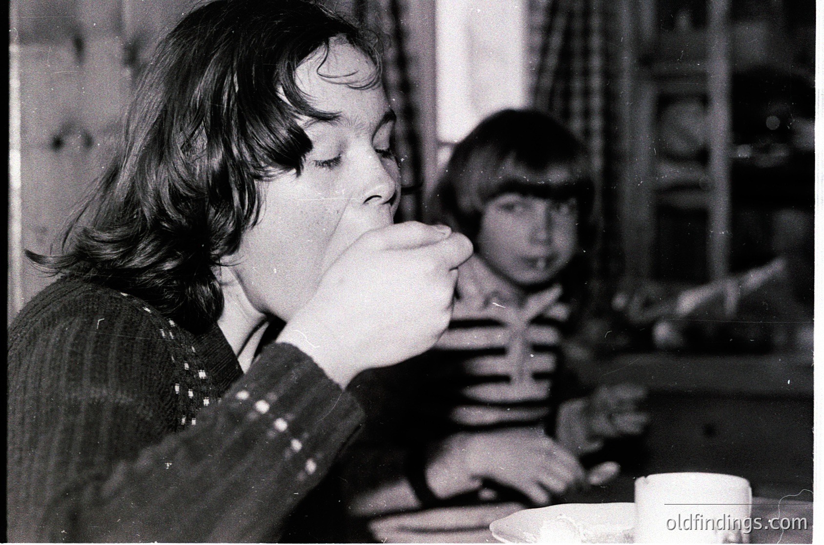 Black-and-white snapshot of two children drinking from cups at a café table, likely mid-20th century. The girl in the foreground wears a patterned sweater, while the boy in the background has short hair and a striped shirt. Wooden table and chairs suggest a vintage European café setting. é