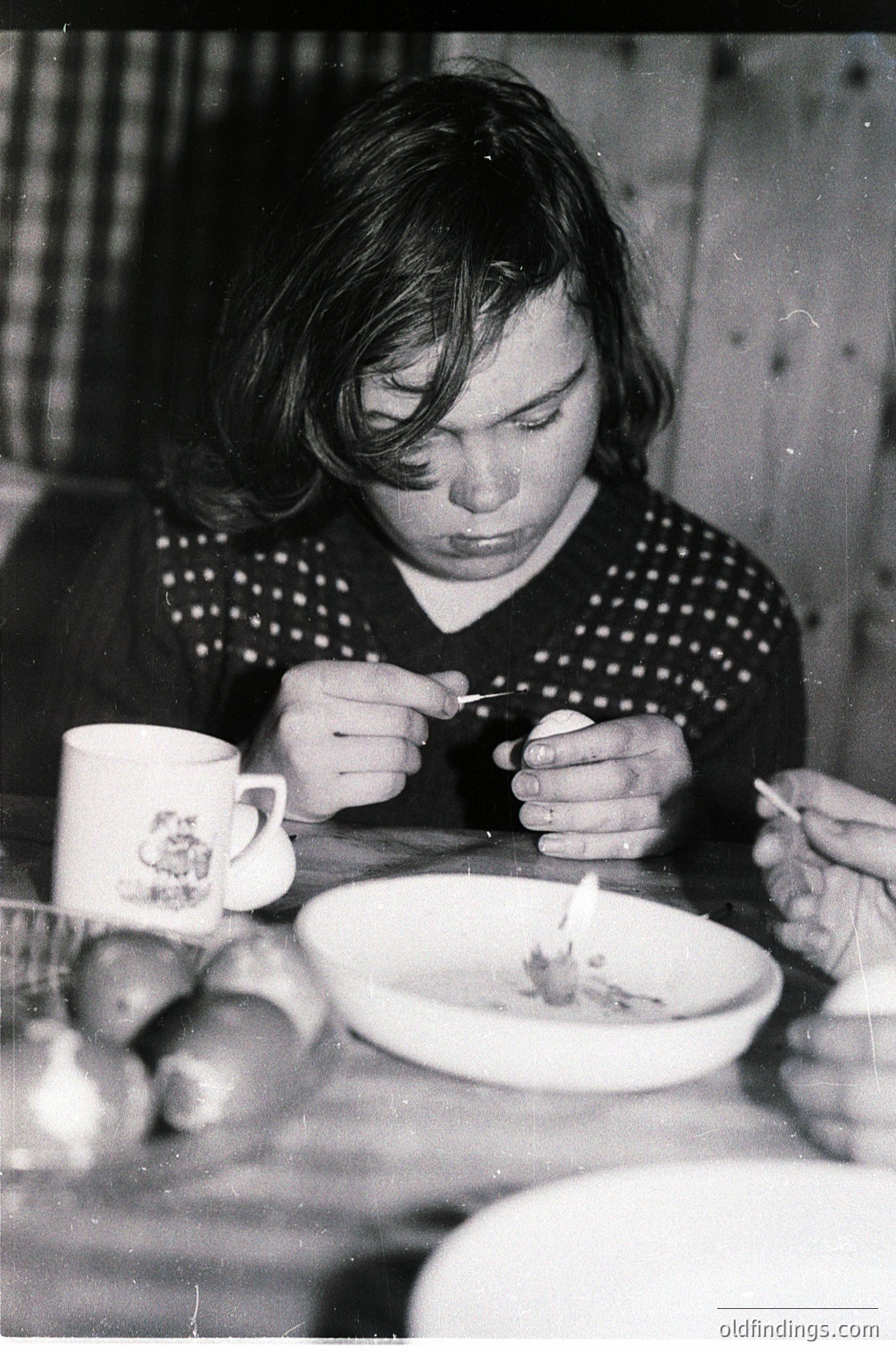 Mid-century domestic scene: Woman in patterned sweater examines food on a plate with a spoon, beside a floral-patterned mug. Wooden table and rustic interior suggest 1950s–1960s home life.