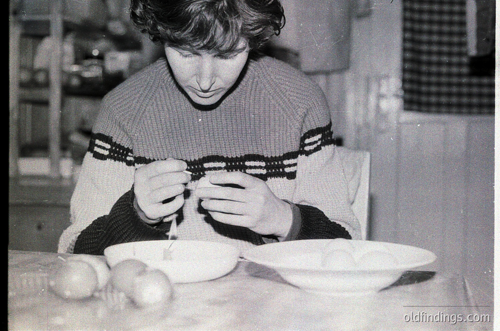 Mid-century kitchen scene featuring a person meticulously peeling a potato with a knife. White plate with peeled potato halves sits beside raw potatoes. Knitted sweater with striped pattern and dark sleeves suggests 1960s–1970s domestic life.