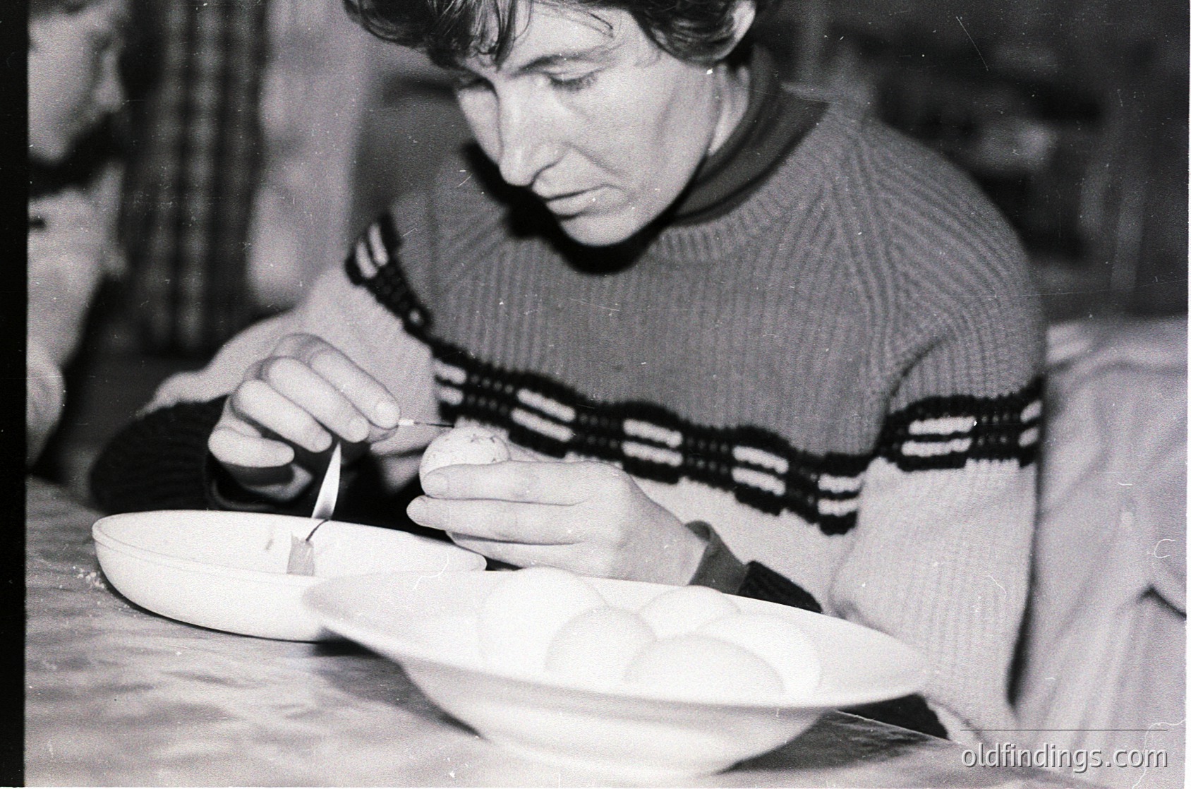 Black-and-white photo of an individual meticulously crafting ceramic pottery using a tool, likely a pottery knife or wire. The scene appears indoors, possibly a workshop or studio, with blurred background suggesting focus on the hands-on process. Sweater with horizontal stripes and a cable-knit pattern indicates mid-20th century fashion. [Mid-century pottery crafting | Handmade ceramic artistry | Studio workspace ]