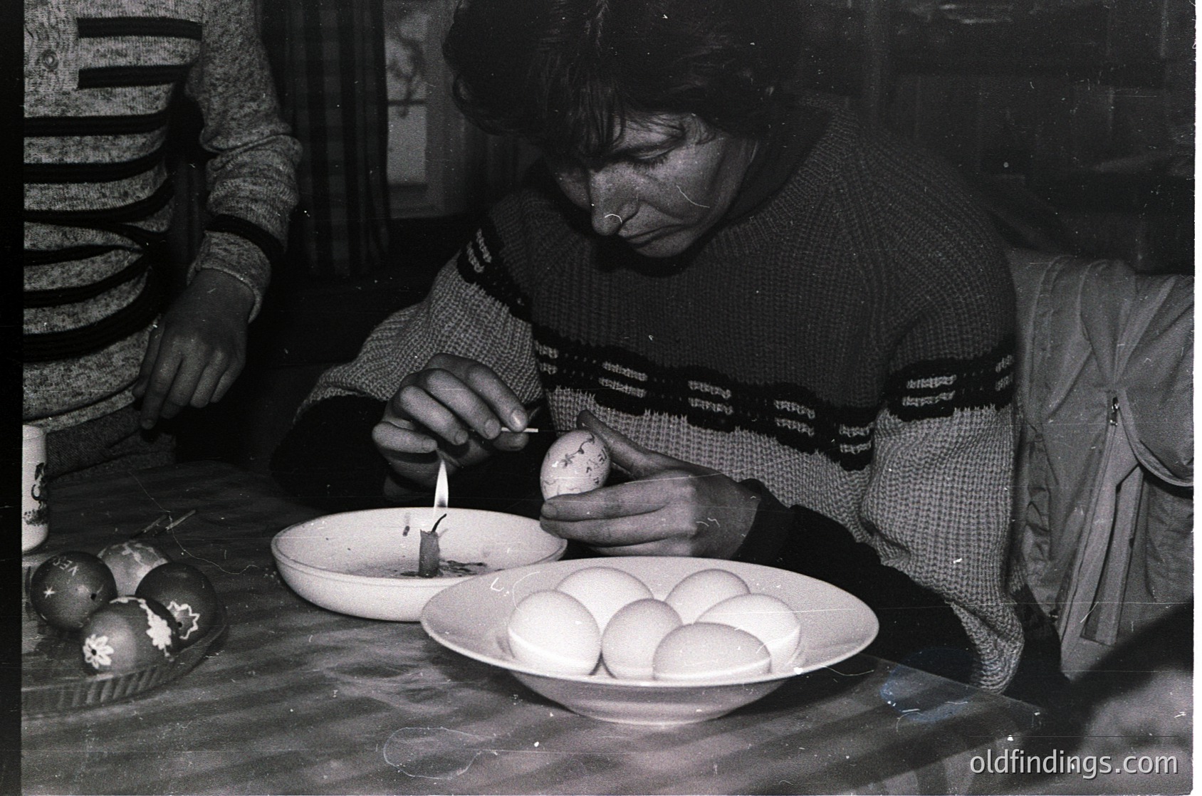 Black-and-white photograph of a woman decorating Easter eggs with a wax pen, seated at a table with a bowl of undyed eggs. She wears a patterned sweater with visible text ("Drapatsy") and a headscarf. Decorated eggs in the background suggest a traditional Easter crafting session. Likely Eastern Europe, mid-20th century.