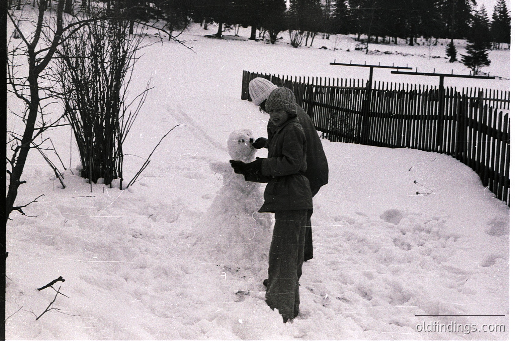 Child sculpting a snowman in a winter garden, mid-20th century. Wooden fence and bare trees frame the scene, suggesting rural or suburban setting. Classic winter attire—scarf, coat, and hat—indicates cold-weather play. Black-and-white composition evokes nostalgia.