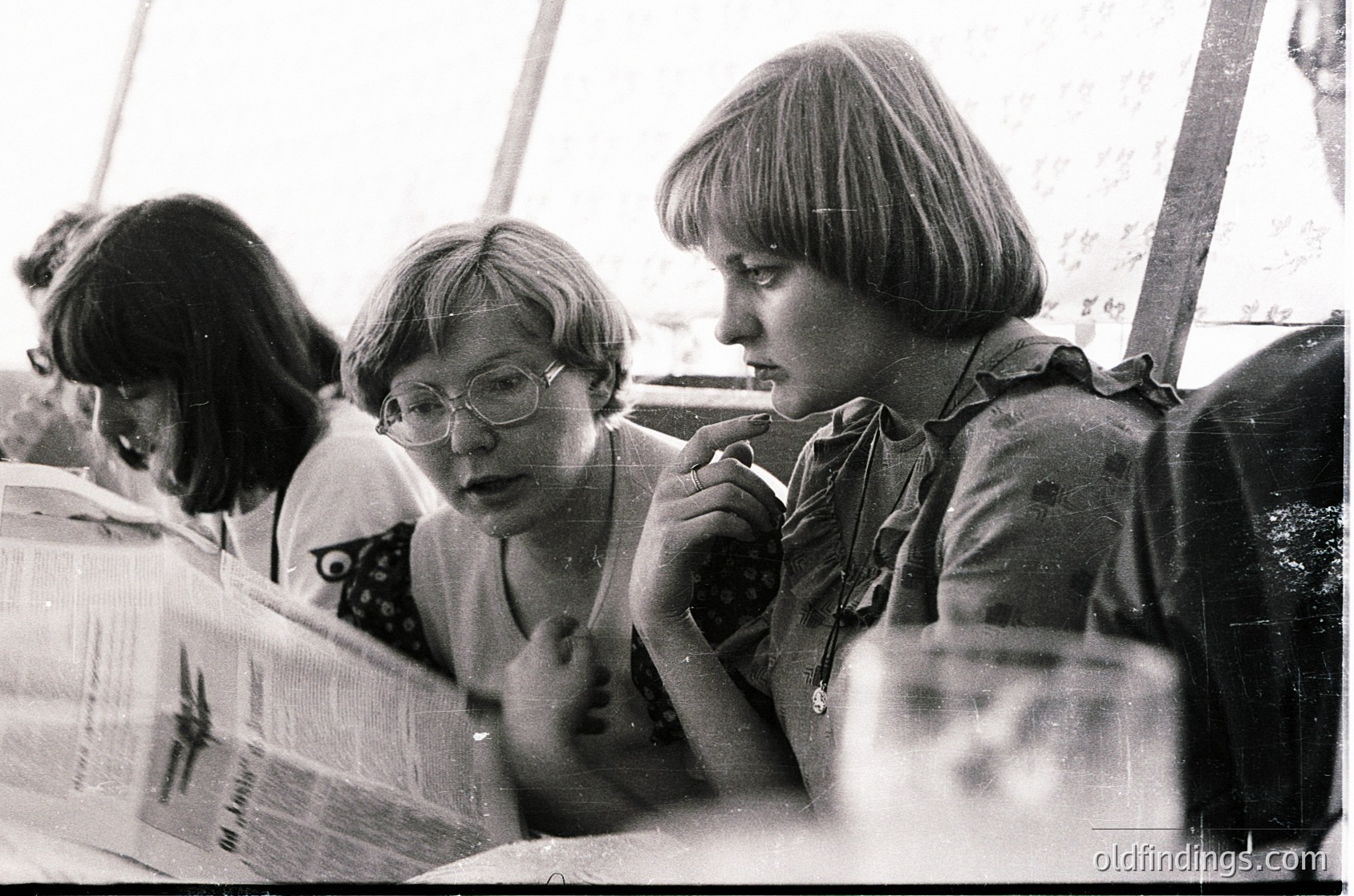 Black-and-white candid shot of three women in casual 1970s attire—short-sleeved blouses, glasses, and shoulder-length hair—engrossed in conversation at a table. Stacked cardboard boxes and a tented structure suggest an outdoor event or festival.