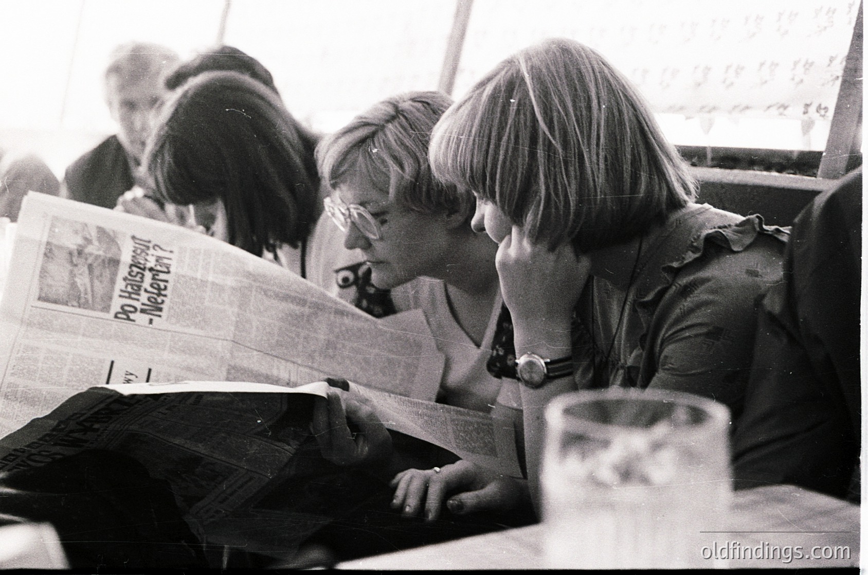 Vintage black-and-white photo of two women engrossed in reading a Dutch-language newspaper (*Po Voetbal-Nieuws*) in a public setting, likely a café or train station. Mid-20th century (1960s–1970s) fashion: round glasses, short hair, and patterned blouses. One woman wears a wristwatch. Glass of beer on the table.