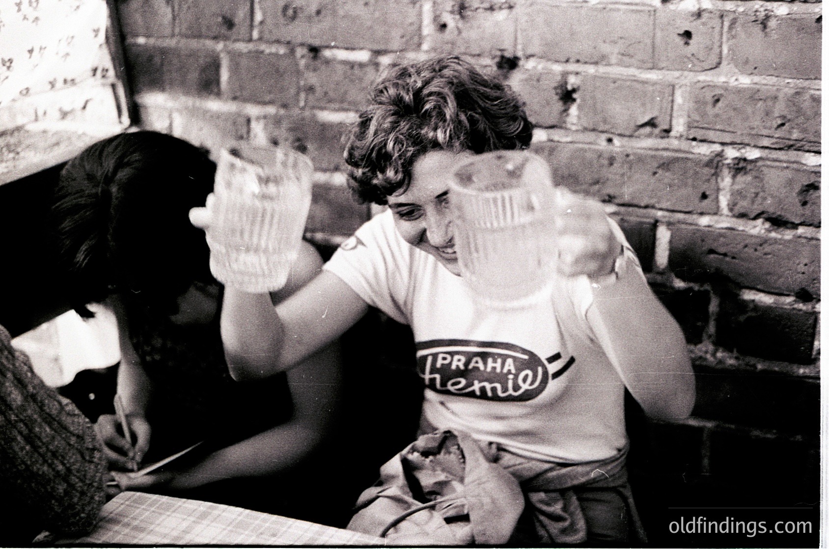 Vintage black-and-white photo of two individuals playfully holding empty glasses in a dimly lit, brick-walled room. The person in the foreground wears a "Praha Pivovary" (Prague Brewery) T-shirt, suggesting a 1960s–1980s Eastern Bloc setting. The scene captures a candid, joyful moment, likely in a social or communal space.