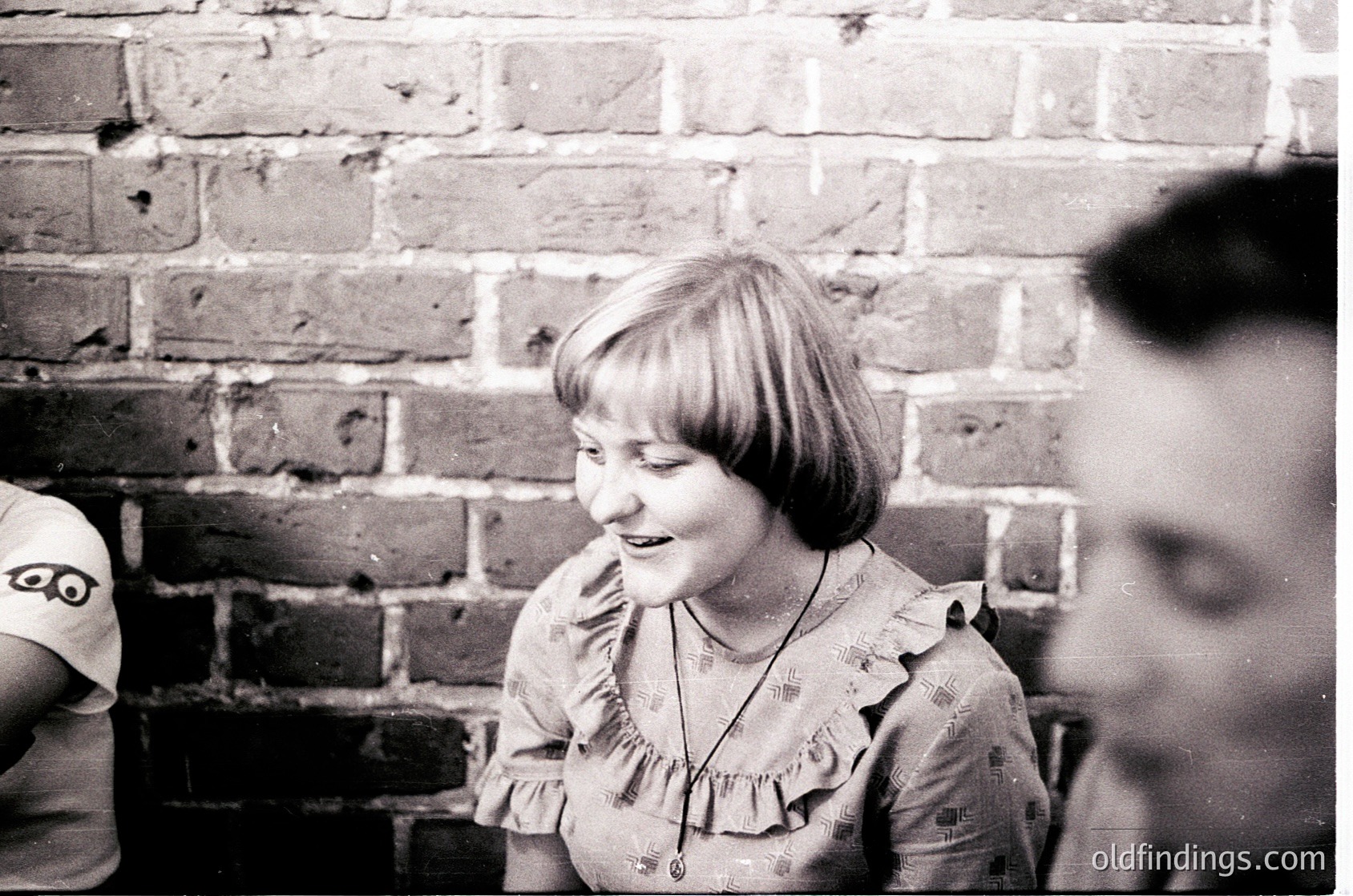 Black-and-white candid shot of a young girl in a ruffled blouse, mid-laugh, against a brick wall.