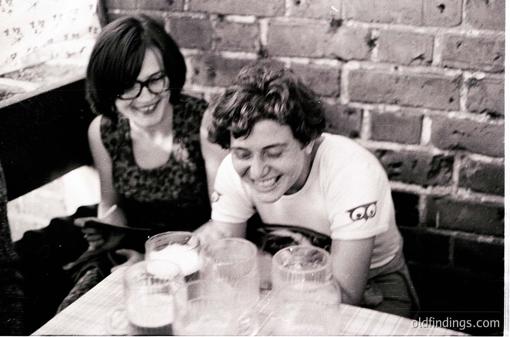 Two individuals share a lighthearted moment over frothy drinks at a rustic brick-walled café, mid-1970s. Glasses, patterned blouse, and vintage T-shirt with embroidered eyes suggest a casual, intimate gathering. éCulture