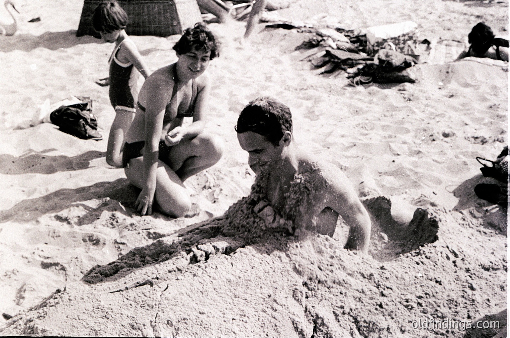 Mid-20th century beach scene: children playing in sand, one crawling while another kneels nearby. Classic 1950s–60s swimwear and cap visible. Crowded shoreline with towels and sandcastles in background.