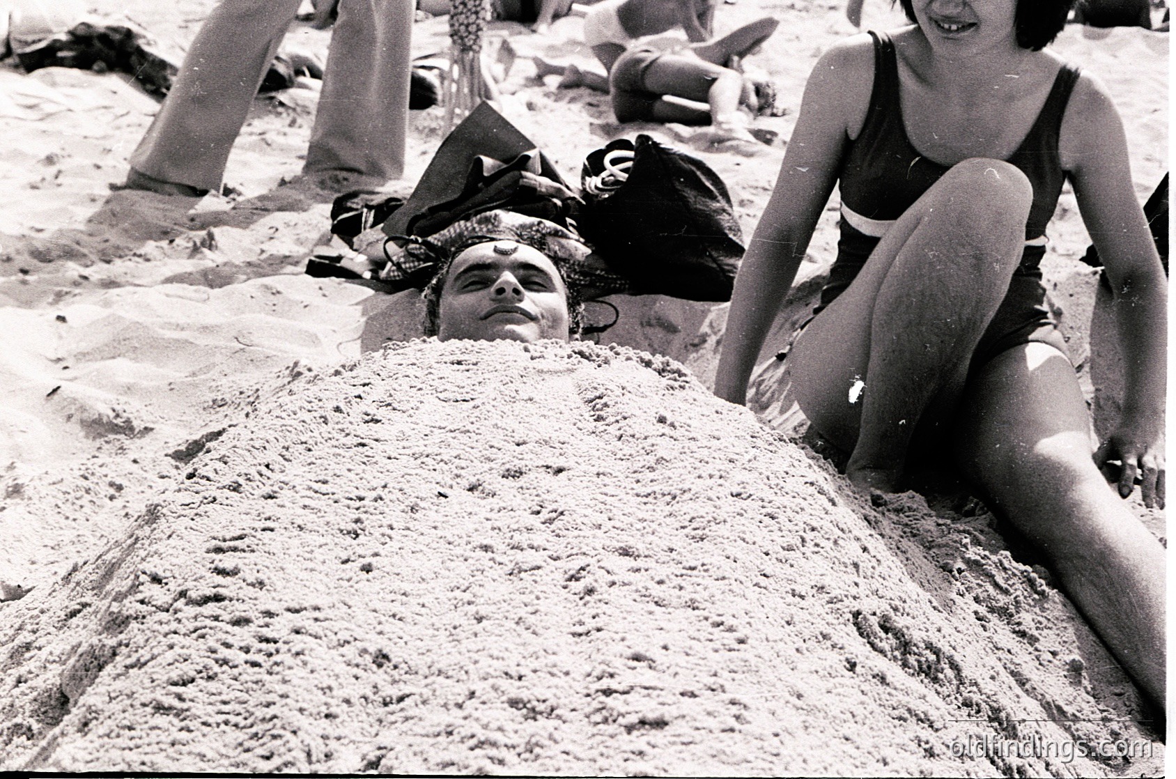 Black-and-white beach scene from the 1960s–70s, featuring relaxed beachgoers in swimwear. A man lies buried in sand up to his chest, while a woman in a bikini sits nearby. Crowded shoreline with others lounging, towels, and footwear scattered. Evokes mid-century beach culture and leisure trends.