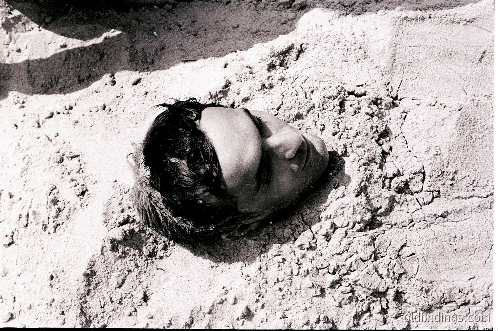 Black-and-white portrait of a person reclining on cracked, textured concrete, likely a rooftop or terrace. The composition highlights the contrast between the smooth skin and rough surface. Style suggests mid-20th century street photography.