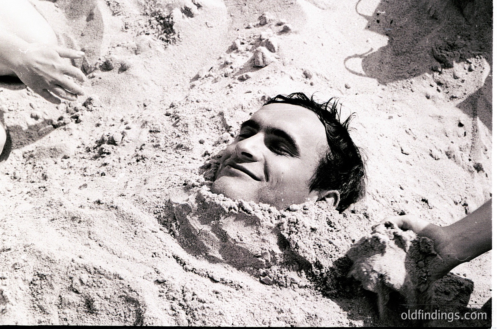 Black-and-white candid of a man buried in sand, partially buried up to his neck, with only his head and shoulders visible. The scene suggests a playful or spontaneous moment, likely at a beach or sandy recreational area. The composition highlights texture and contrast between light and dark tones.