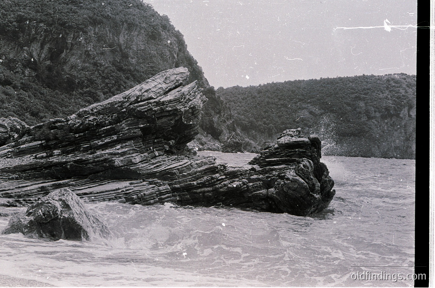 Vintage black-and-white coastal scene featuring jagged, stratified rock formations jutting from turbulent waters. Dense forest covers the cliffside background, suggesting a rugged, untouched landscape. Likely mid-20th century due to grainy texture and monochrome.