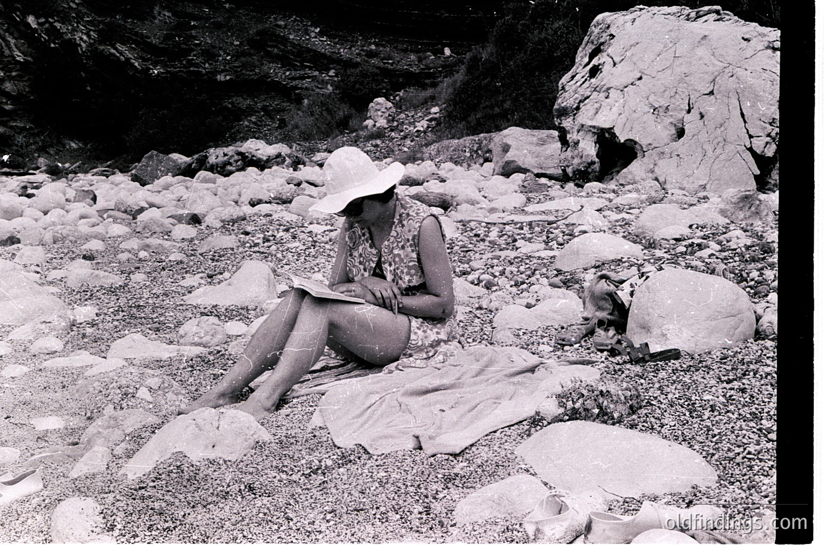Mid-century outdoor scene: Woman in wide-brimmed hat and sleeveless dress reads on a blanket amid rocky terrain, likely a coastal or mountainous area. Style suggests 1950s–1960s leisure photography.