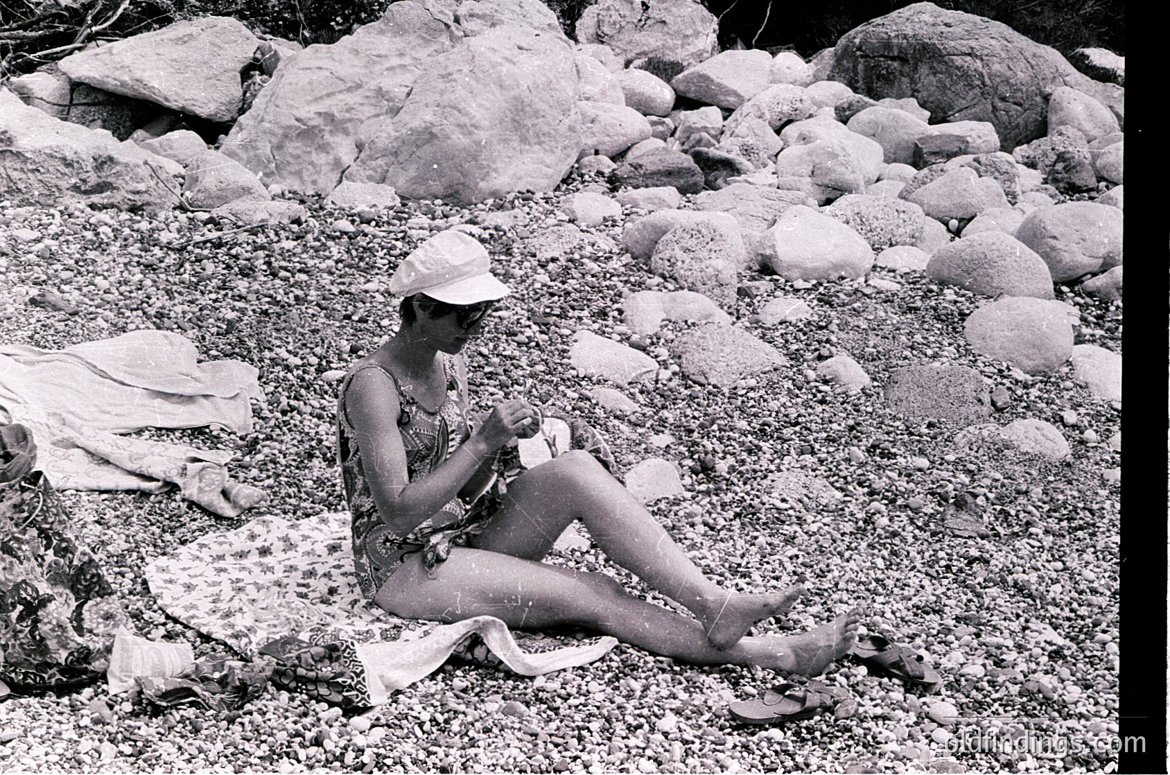Mid-century beach scene featuring a woman in a floral swimsuit and straw hat, seated on a towel among smooth river rocks. Her relaxed posture suggests a leisurely 1950s–1960s outdoor setting, possibly a lakeside or riverside retreat.