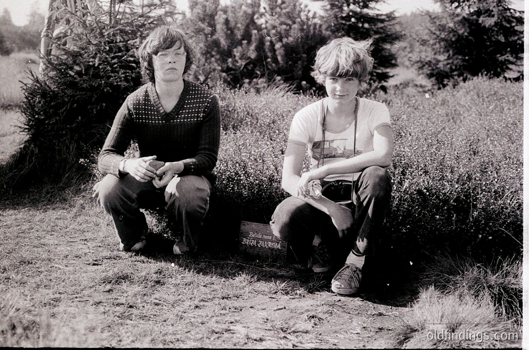 Two individuals pose outdoors on grass beside a gravestone with Hindi script, likely indicating a memorial. The man wears a patterned sweater and dark pants; the boy sports a graphic tee and rolled cuffs. Blurred greenery and trees frame the scene, suggesting a rural or park setting. Black-and-white aesthetic suggests vintage or archival quality, possibly late 20th century. Ideal for historical research or cultural documentation.