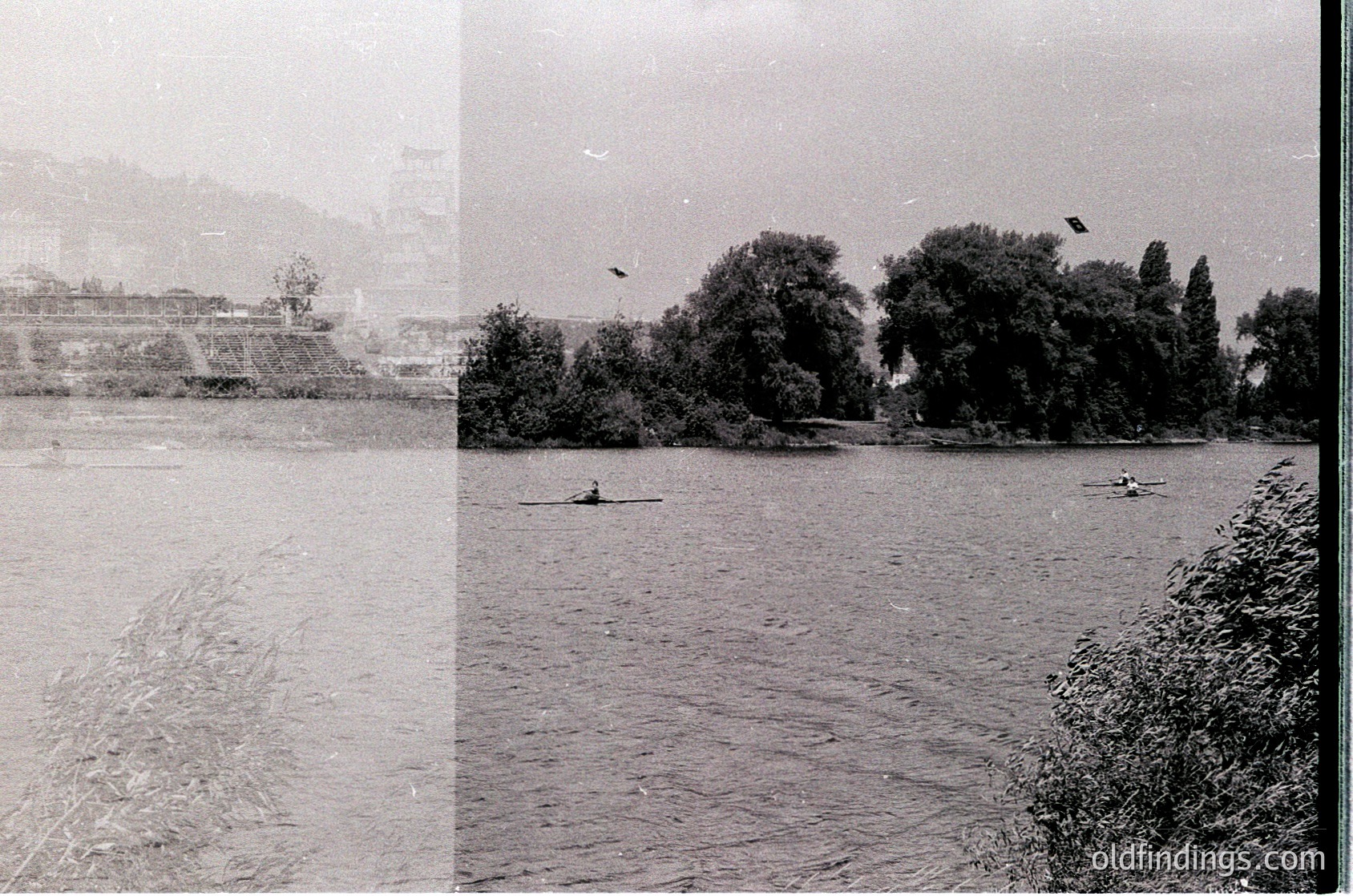 Vintage black-and-white shot of a calm river scene with two canoeists paddling. Dense foliage lines the right bank, while a distant industrial structure and hilly terrain appear on the left. Overcast skies and muted tones suggest mid-20th century. Ideal for historical or nature-themed stock.