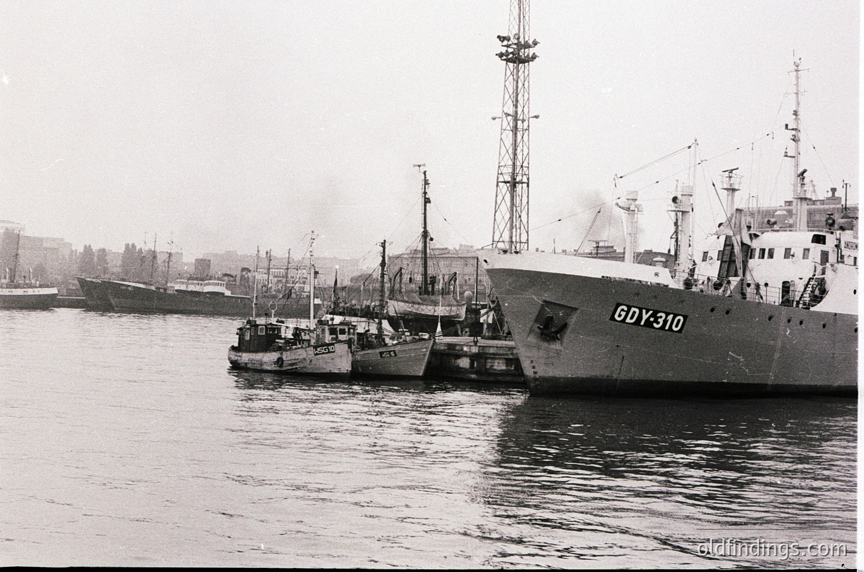 Vintage black-and-white port scene featuring three industrial fishing vessels docked side-by-side. The lead ship, marked "GDY-310," displays Soviet-era design with a tall mast and smokestack. Smaller trawlers and a distant industrial ship frame the composition. Urban skyline with low-rise buildings in background suggests a coastal city port, likely Eastern Bloc, 1960s-70s.