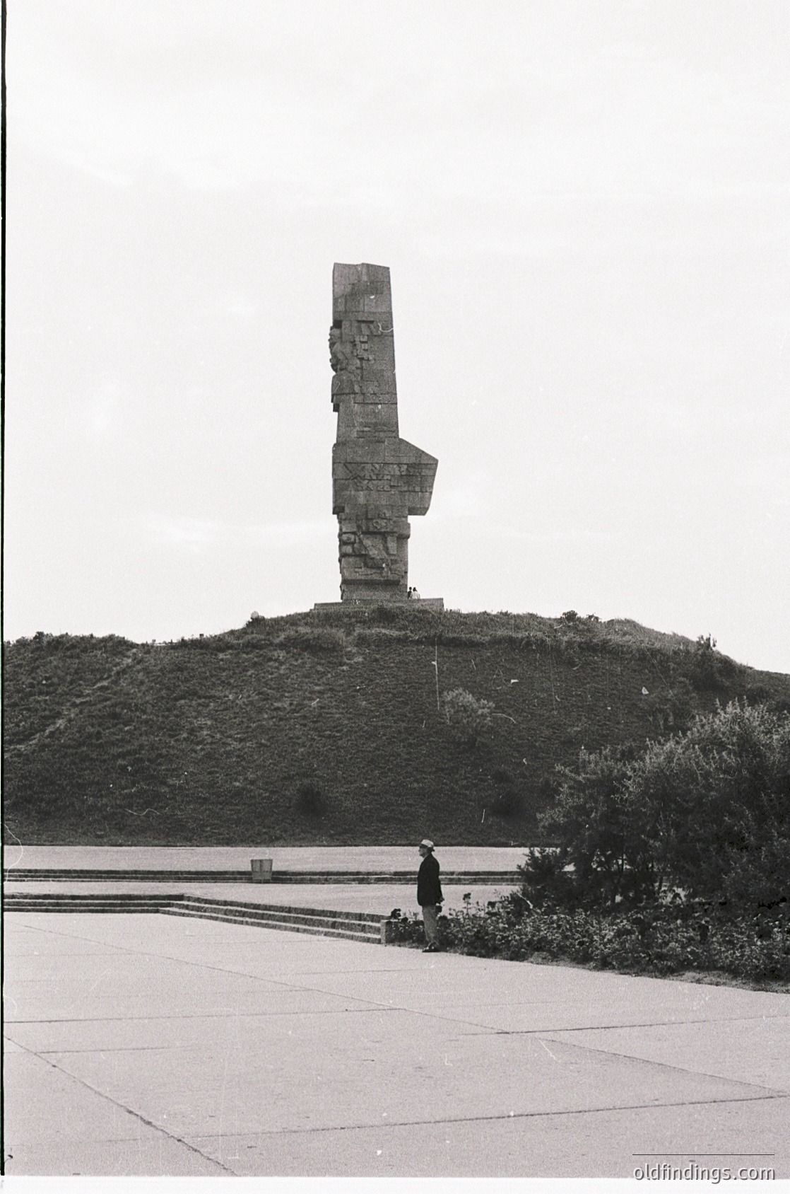 Monumental concrete slab with a raised "V" symbol atop a grassy hill, likely a Soviet-era victory memorial. Two individuals in mid-20th-century attire pose near the base. Overcast sky and paved pathway suggest a public park setting.