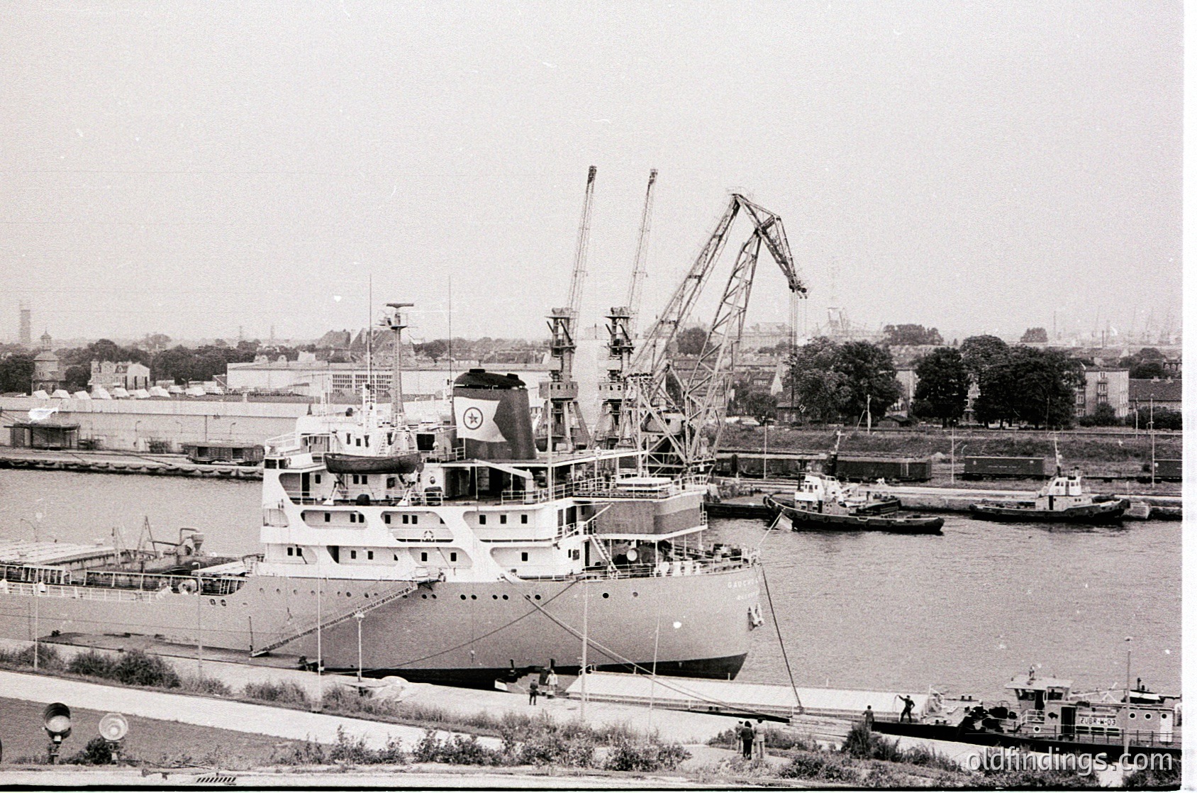 Mid-20th century industrial port scene featuring a large cargo ship docked alongside cranes and smaller vessels. Distinctive Soviet-era maritime flag on the ship’s stern. Urban backdrop with warehouses and greenery along the waterfront.