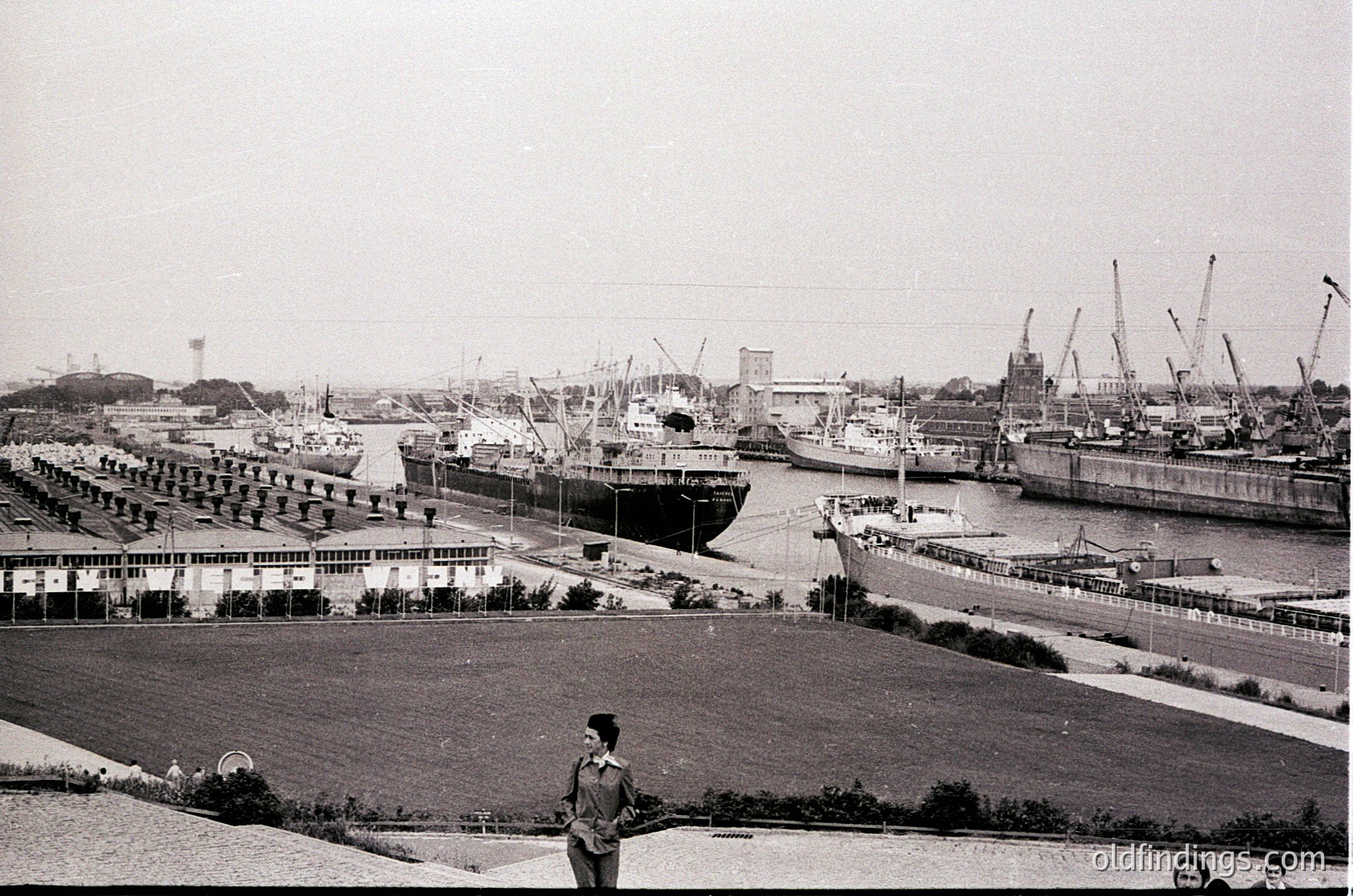 Black-and-white port scene featuring mid-20th century maritime infrastructure. Docked cargo ships, cranes, and industrial buildings line a bustling harbor. Prominent low-rise concrete structures with uniform windows flank the waterfront. A lone figure in a suit stands on a grassy embankment, adding scale to the industrial landscape.