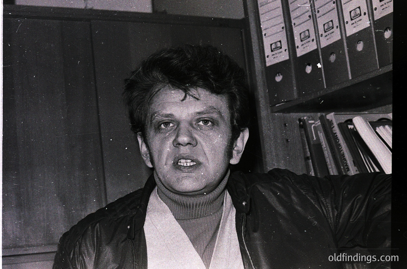 Black-and-white portrait of a man in a turtleneck sweater and leather jacket, standing indoors near stacked audio cassettes and bookshelves. Mid-20th century studio lighting and retro styling suggest a 1970s–1980s music or media setting.