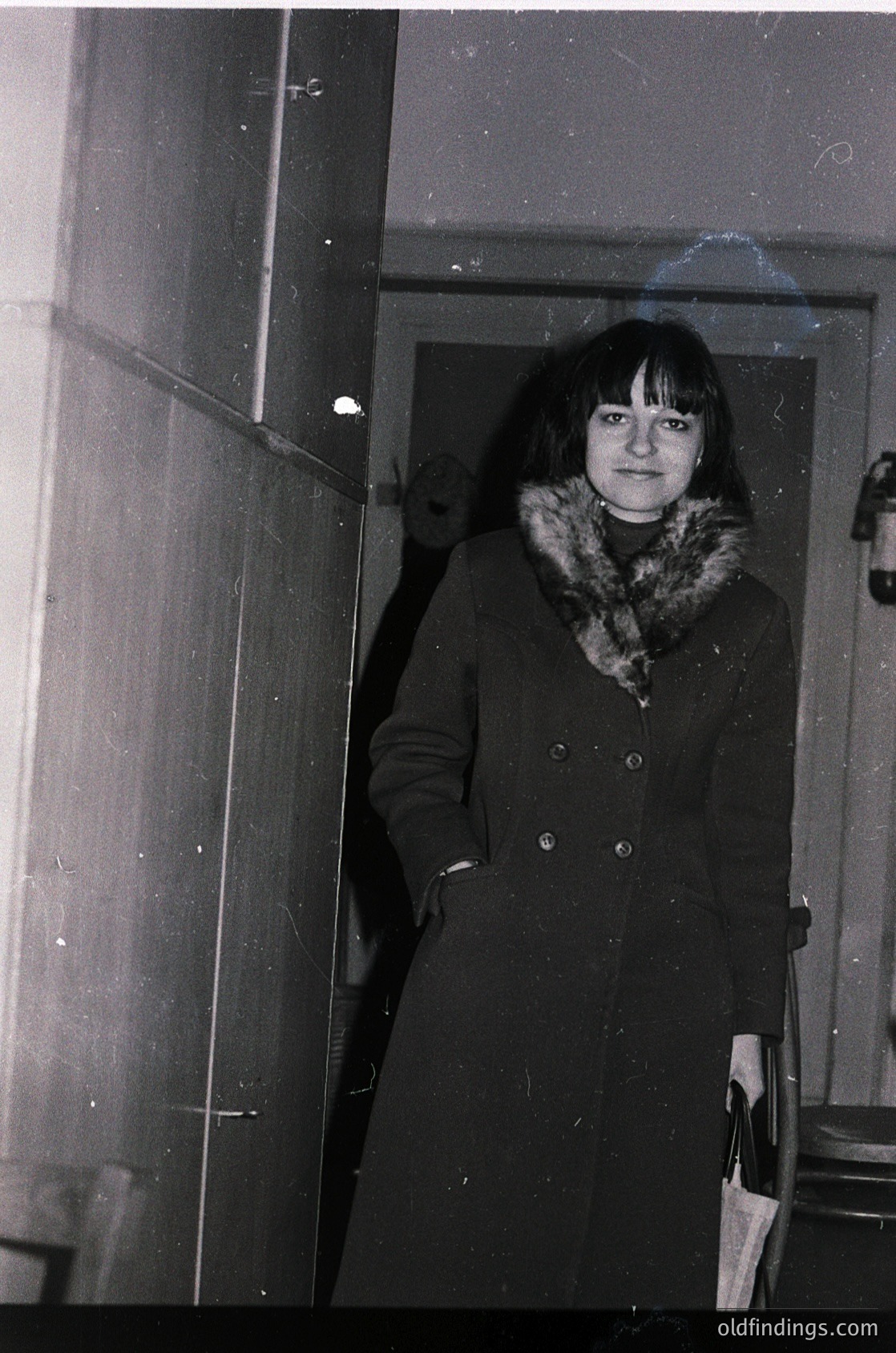 Black-and-white portrait of a woman in a long fur-collared coat, standing indoors near a frosted glass door. The coat’s buttoned cuffs and structured silhouette suggest 1960s–1970s fashion. The blurred background hints at a residential or institutional hallway.