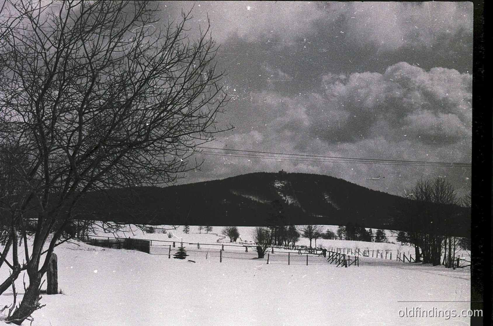 Mid-century black-and-white winter landscape featuring snow-covered fields, barbed-wire fencing, and a distant hilltop structure. Dramatic clouds and bare trees frame the scene, evoking 1950s–1960s rural isolation.