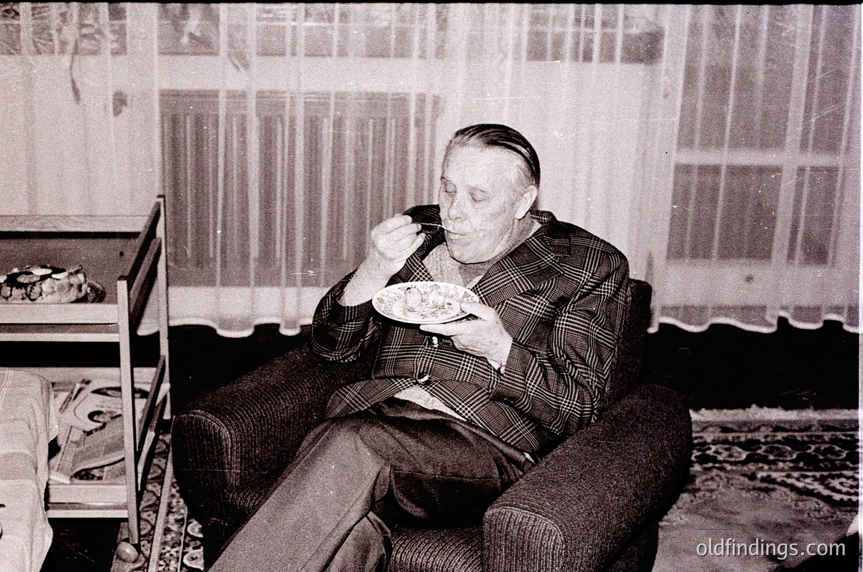 Mid-century man in plaid suit eats from a bowl while seated in a vintage armchair, suggesting 1950s–1960s domestic life. Curtains, patterned rug, and a small table with a cake hint at mid-20th-century interior design.