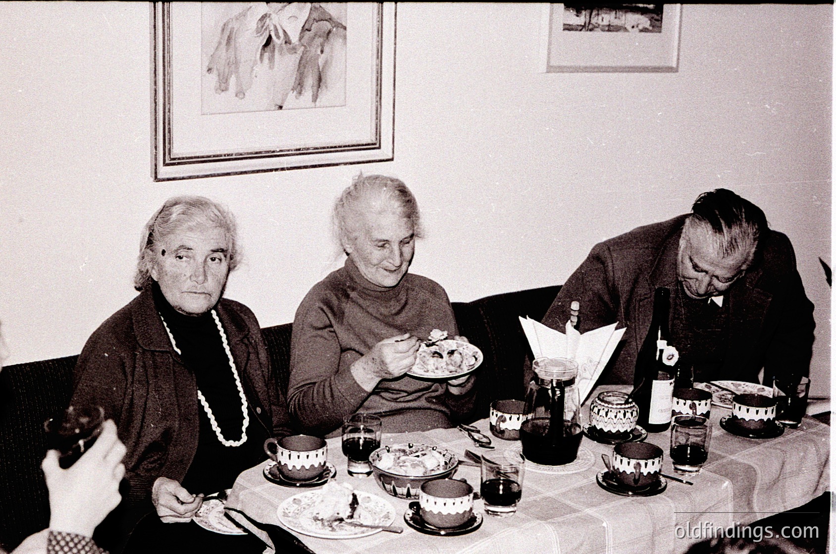 Three elderly individuals seated around a table with vintage tableware—teacups, plates, and a bottle—suggesting a mid-20th century gathering. Plain wall art and neutral tones indicate a modest indoor setting, likely a private home or community event. Style and lighting hint at 1950s–1960s era.