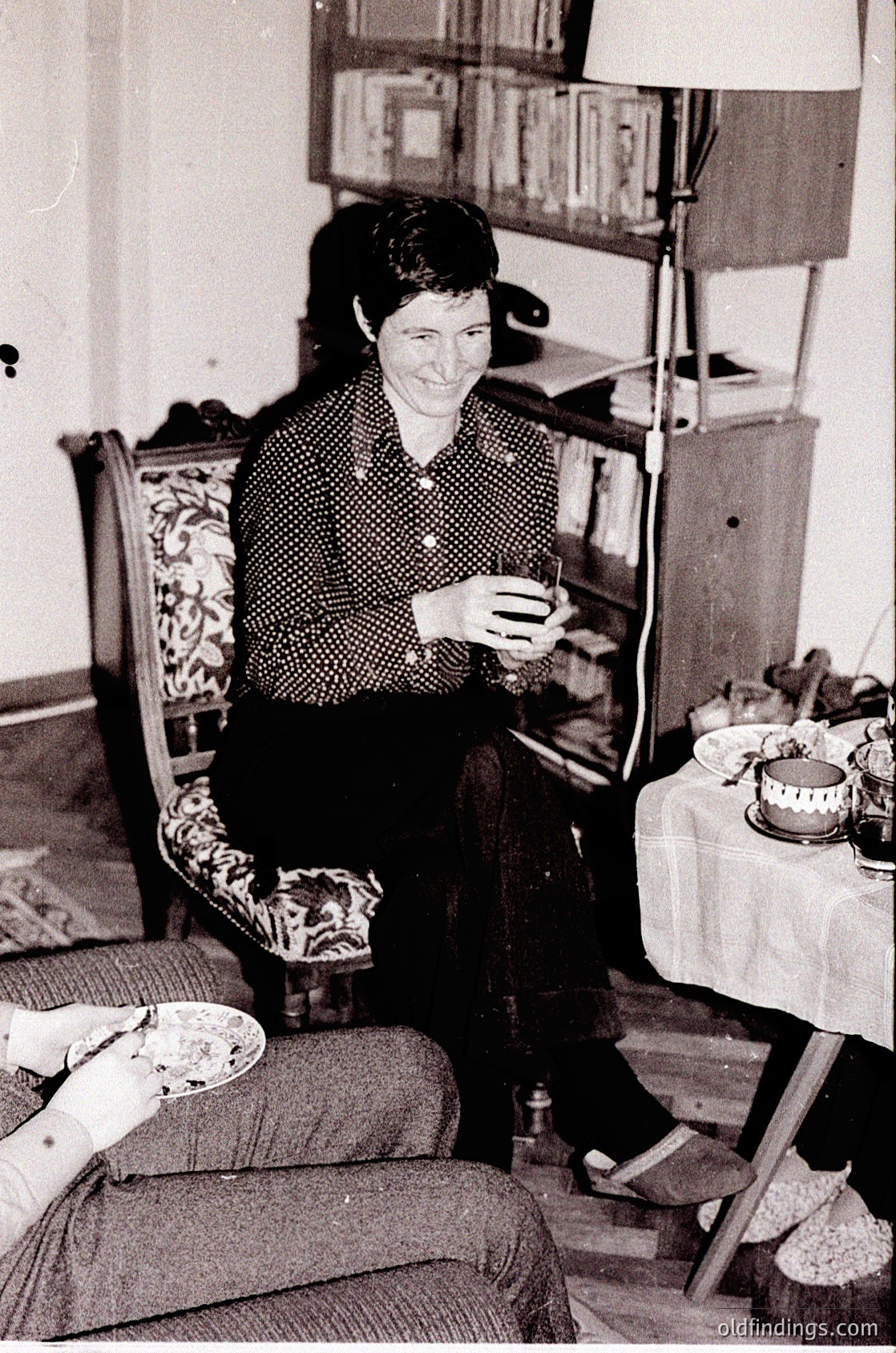 Vintage black-and-white interior shot of a woman seated on a patterned sofa, holding a small object (likely a cigarette or pipe) in hand. Surrounding her: a wooden table with crockery, a bookshelf with books, and a chair with floral upholstery. Mid-20th century domestic setting, suggestive of European or Eastern European style.