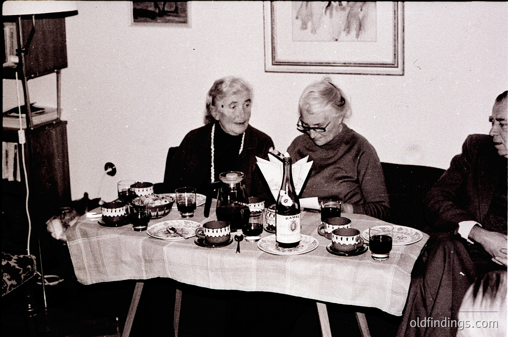 Three elderly women seated around a table set with vintage glassware, plates, and a bottle of wine, likely mid-20th century. Plain tablecloth, framed artwork on wall, and simple interior suggest a modest domestic setting.
