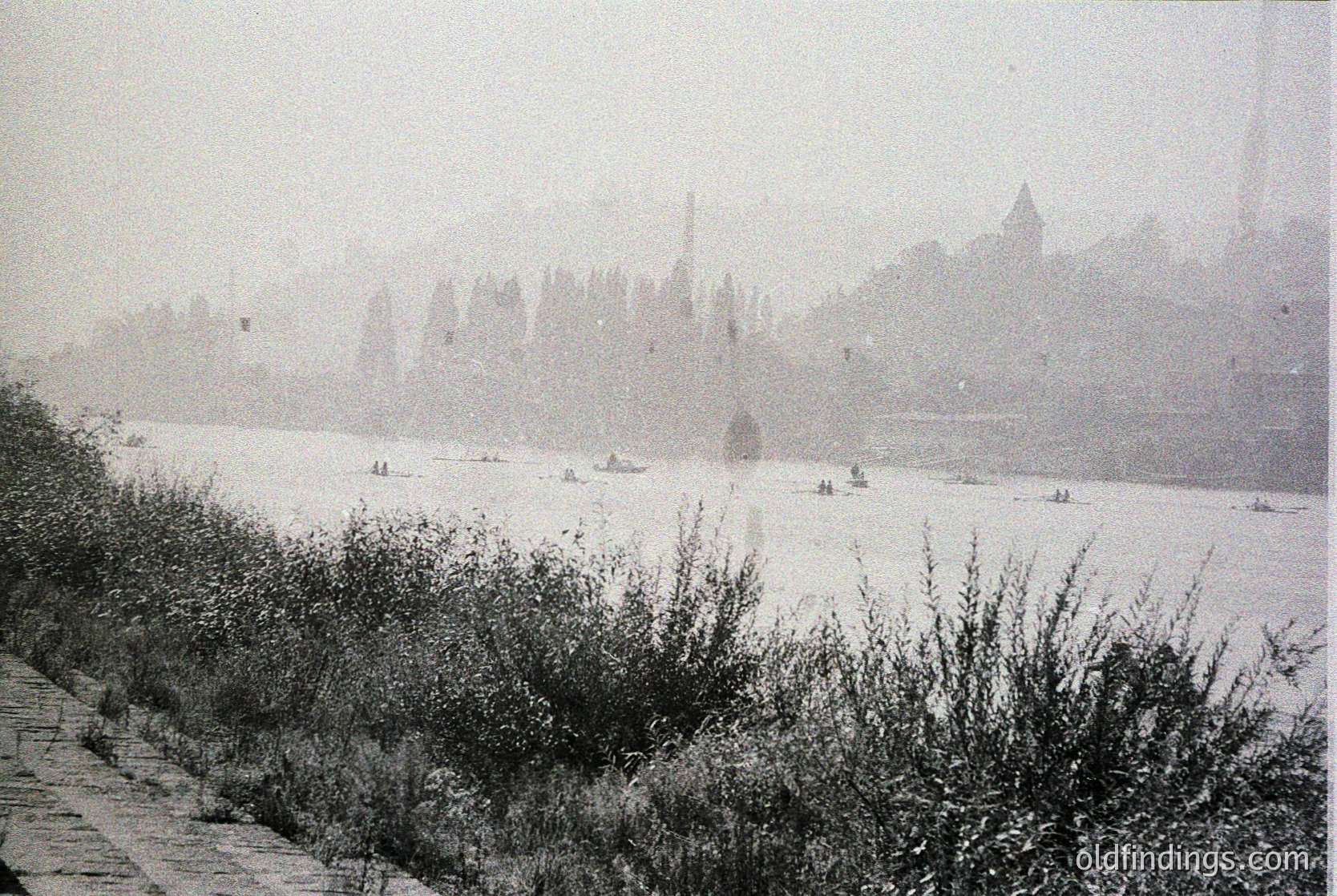 Vintage black-and-white river scene with misty castle ruins on distant hillside, likely European. Small boats dot the water, framed by overgrown vegetation and a stone path. Fog obscures details, adding timeless atmosphere. Potential --- *Note: Exact location/time indeterminate, but stylistic cues suggest 19th-century European riverine setting.*