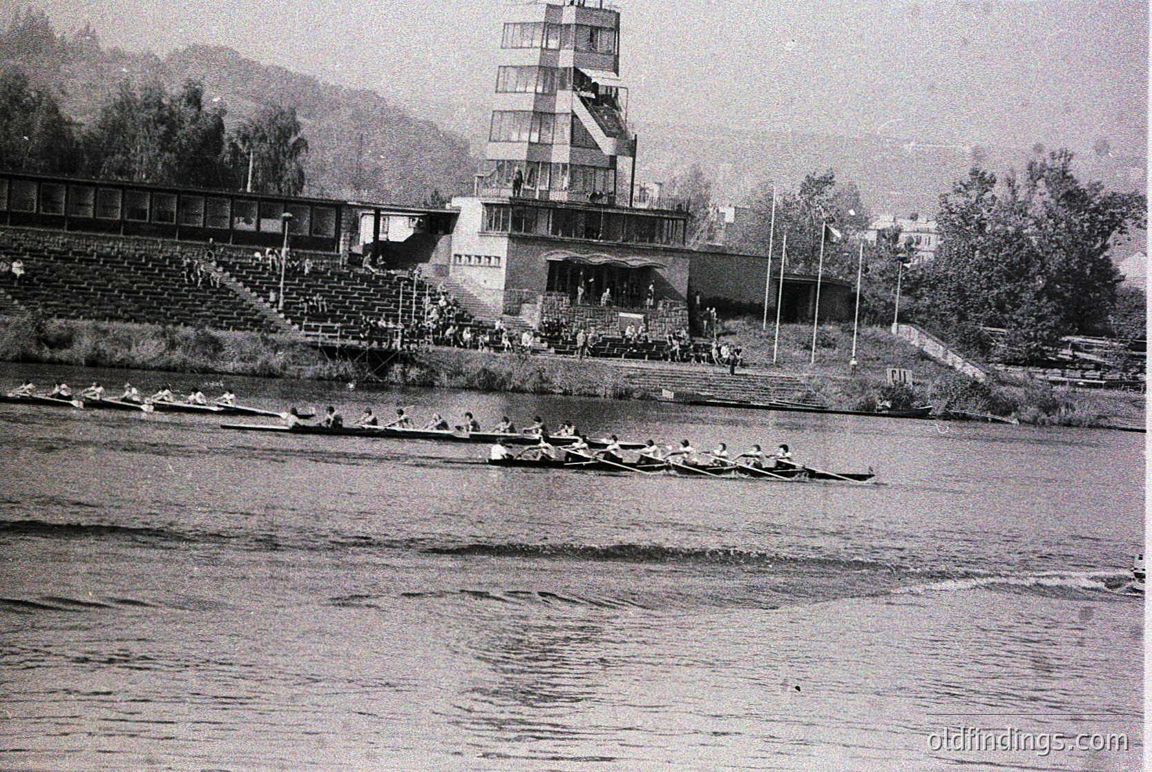 Black-and-white shot of a rowing competition at a stadium-style lakeside venue, featuring tiered concrete seating and a multi-level control tower. Eight-person shells race on calm waters, with spectators lining the banks. Architectural Brutalist elements dominate the background. Likely Eastern Bloc-era, 1960s–1970s.