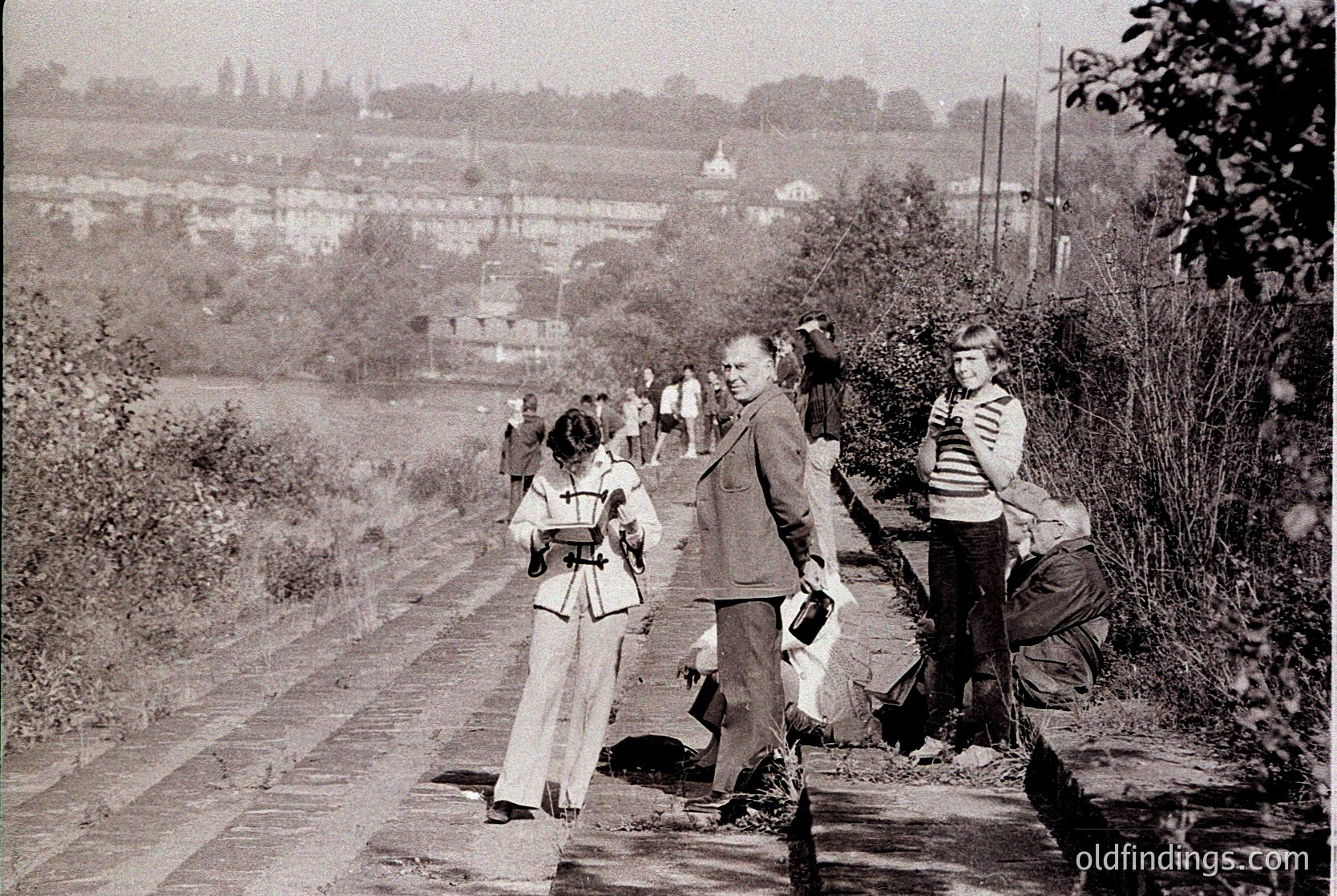 Group of people in mid-20th-century attire clearing a rural pathway, likely post-WWII. Distinctive 1950s-60s clothing: men in suits, women in long skirts. Background shows modest brick houses and a distant minaret, suggesting Eastern European setting.