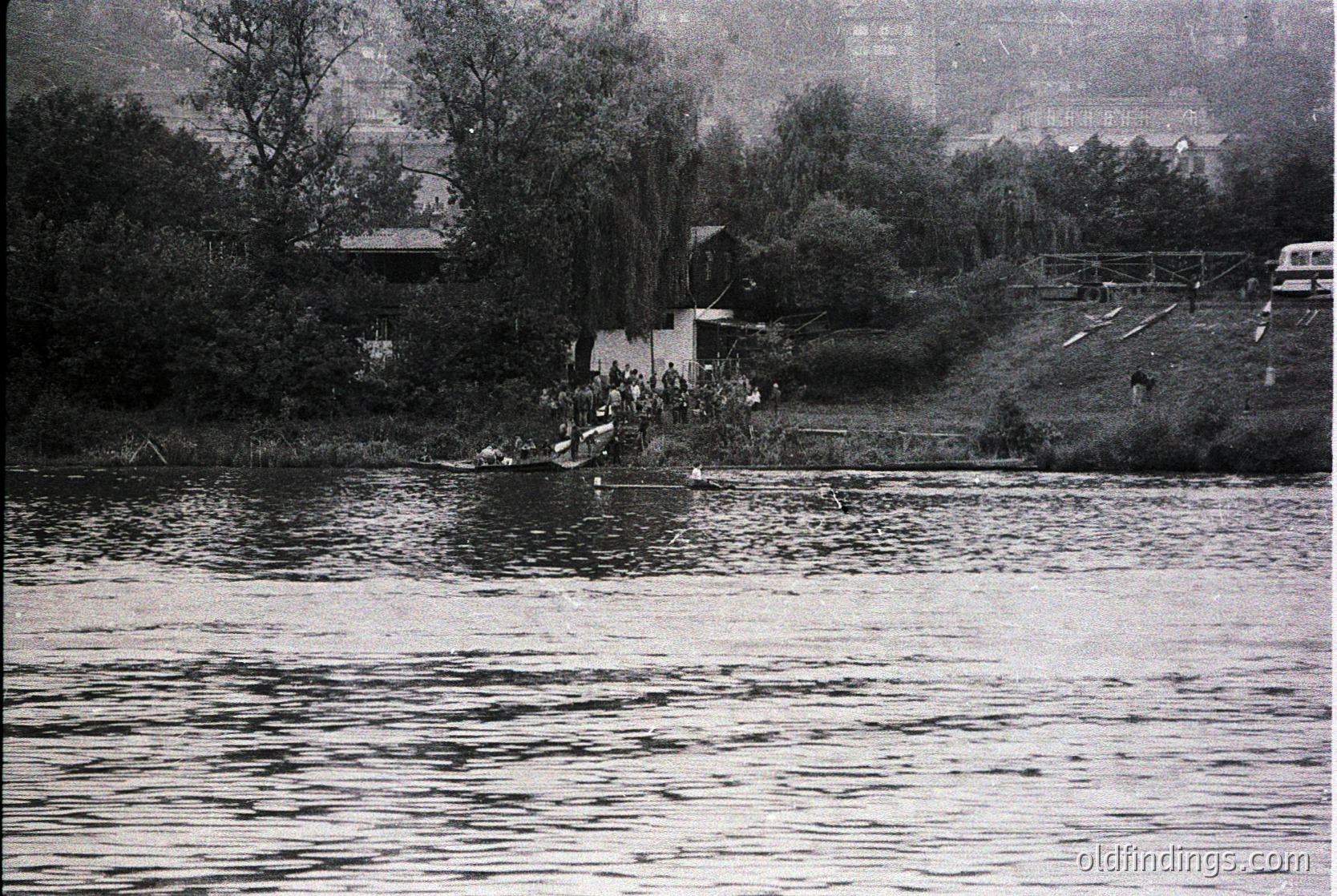 Black-and-white shot of a crowded riverside gathering, likely mid-20th century. A dense group of people stands on a rocky shore, some wading into shallow water. A lone figure in a boat or raft floats nearby. Dense foliage and a distant bridge frame the scene. Urban skyline visible in background.