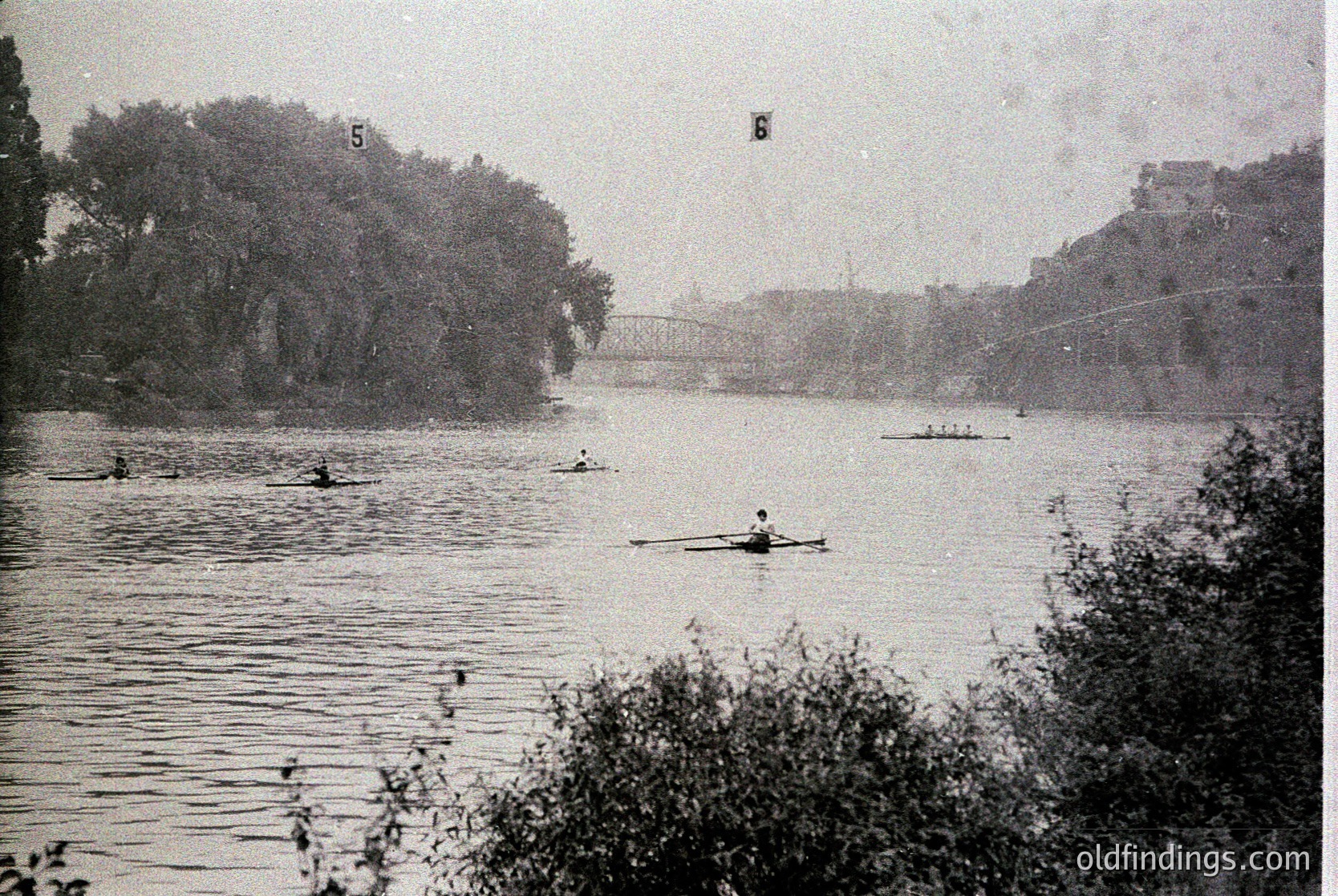 Vintage sepia-toned rower on calm river with bridge and trees in background, likely early 20th century. Wooden sculling boat and lone rower in motion, surrounded by other watercraft. Urban park or city waterway setting.