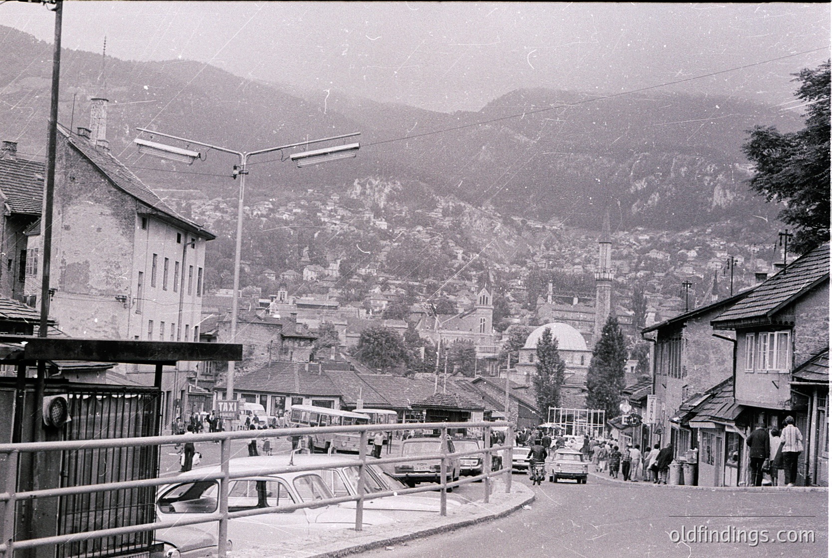 Black-and-white street scene in a mountainous European town, likely –70s. Narrow road flanked by parked vintage cars, pedestrians, and modest buildings with sloped roofs. Distant mosque/minaret and domed church suggest mixed religious architecture. Rugged alpine backdrop with misty hills.