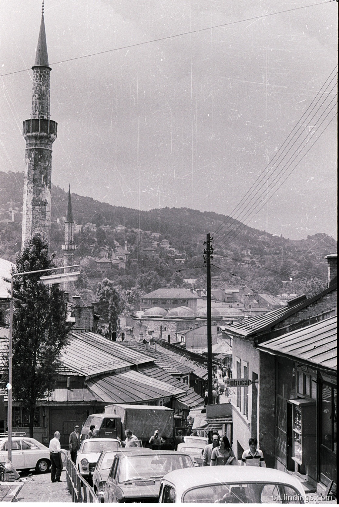 Vintage black-and-white street scene featuring a prominent Ottoman-style minaret and dense urban rooftops. Mid-20th century cars and pedestrians populate a hilly, Mediterranean town. --- *Note: Exact location indeterminate but stylistic cues suggest Southeastern Europe (e.g., Balkans).*