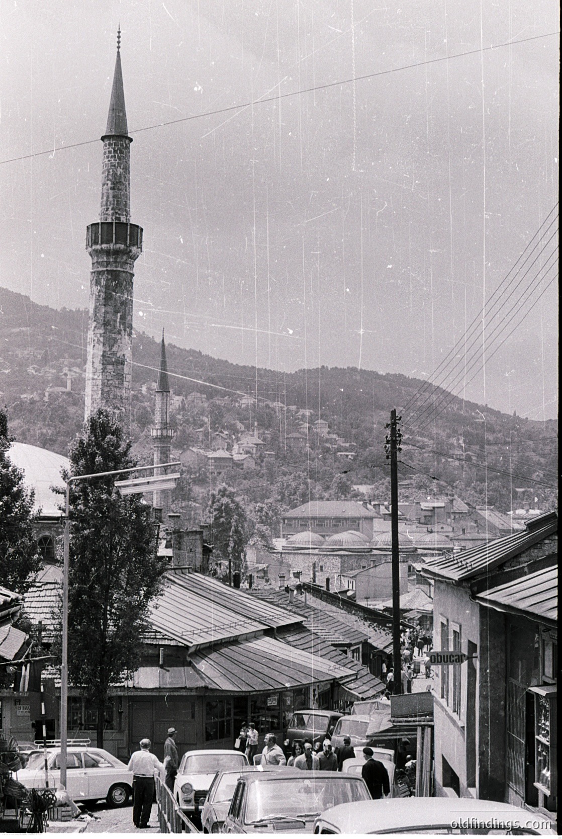 Black-and-white street scene featuring Ottoman-style minarets and domes in a hilly town, likely or . Mid-20th century architecture with vintage cars and pedestrians. Urban landscape with mixed residential/commercial buildings.