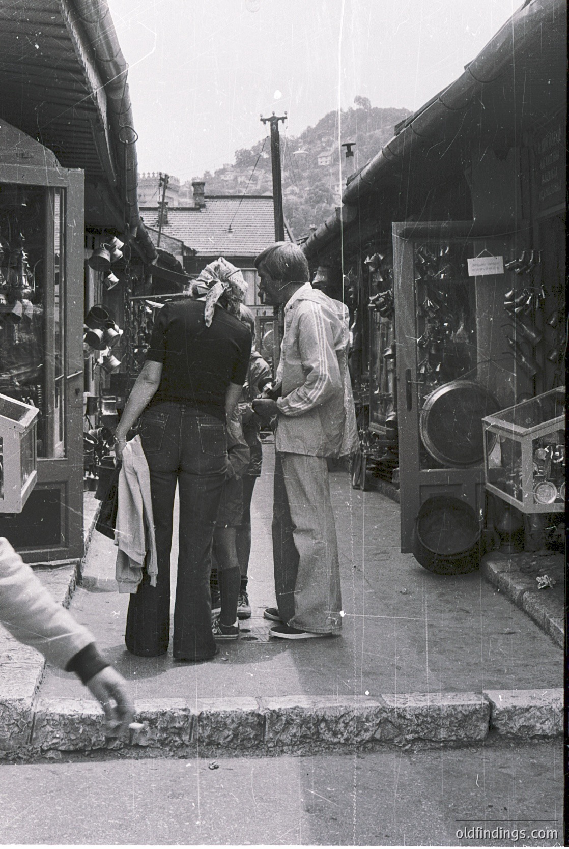 Vintage street scene featuring two men examining a large, vintage mechanical device (likely a sewing machine) in a narrow alleyway lined with shops. Wooden storefronts display textiles and household goods. The setting suggests a 1960s–1970s European market town, possibly or . Blurred motion indicates candid, documentary-style photography.