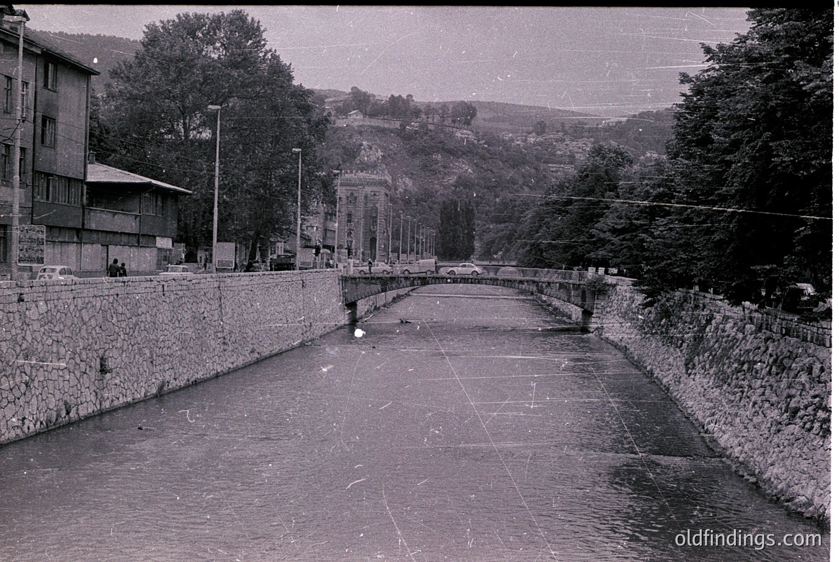 Mid-20th century urban canal bridge with stonework and pedestrian pathway, flanked by Soviet-era concrete buildings. Overhead wires and streetlamps suggest planned infrastructure.