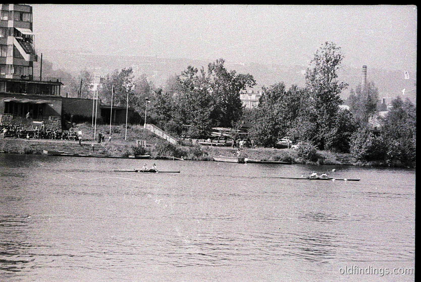 Black-and-white shot of a riverbank scene with rowers practicing in the mid-20th century. Dense greenery frames a rowing lane, with multiple boats and paddlers in sync. Industrial buildings and a smokestack hint at urban proximity. Likely a training session or club activity.