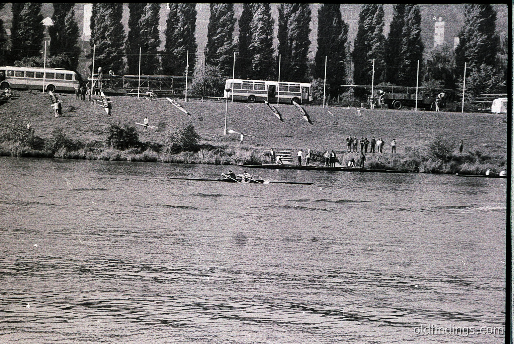 Vintage black-and-white shot of a lakeside scene with mid-20th century buses parked on grassy embankment. People swim, wade, and gather near the water’s edge, while others stand or sit on the shore. Dense pine trees frame the background, and a distant building is visible. Likely a public recreational area from the or .