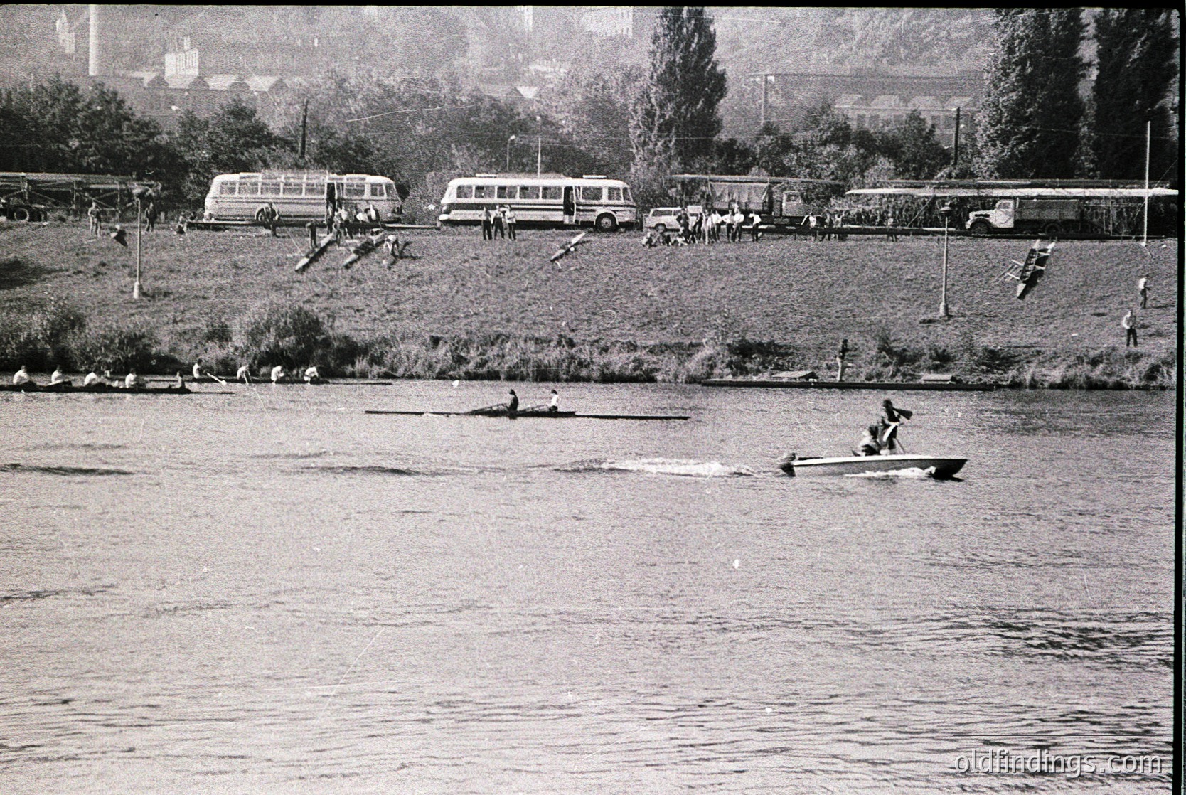 Vintage black-and-white scene of rowers competing on a calm river, with vintage buses and spectators lining the shore. Mid-20th century European rowing event, likely 1950s–1960s.
