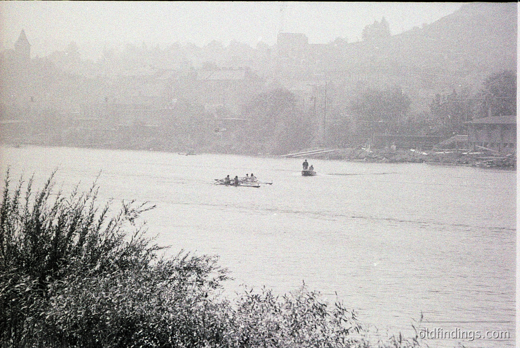 Vintage black-and-white river scene with misty urban backdrop. Small rowboats and a motorboat navigate calm waters, framed by reeds. Historic European architecture—likely a castle or fortified town—fades into haze. Mid-20th century recreational activity.