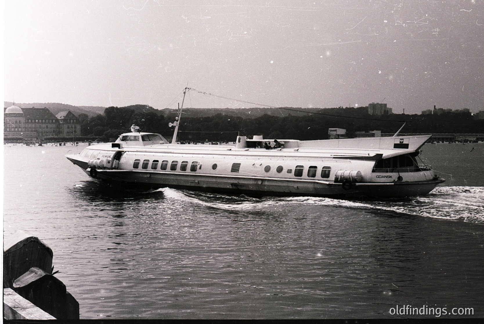 Mid-20th century ferry with sleek, elongated hull and circular portholes cruising on calm waters. Urban shoreline with brick buildings and greenery in background. Likely Eastern European coastal city, 1950s–1970s.