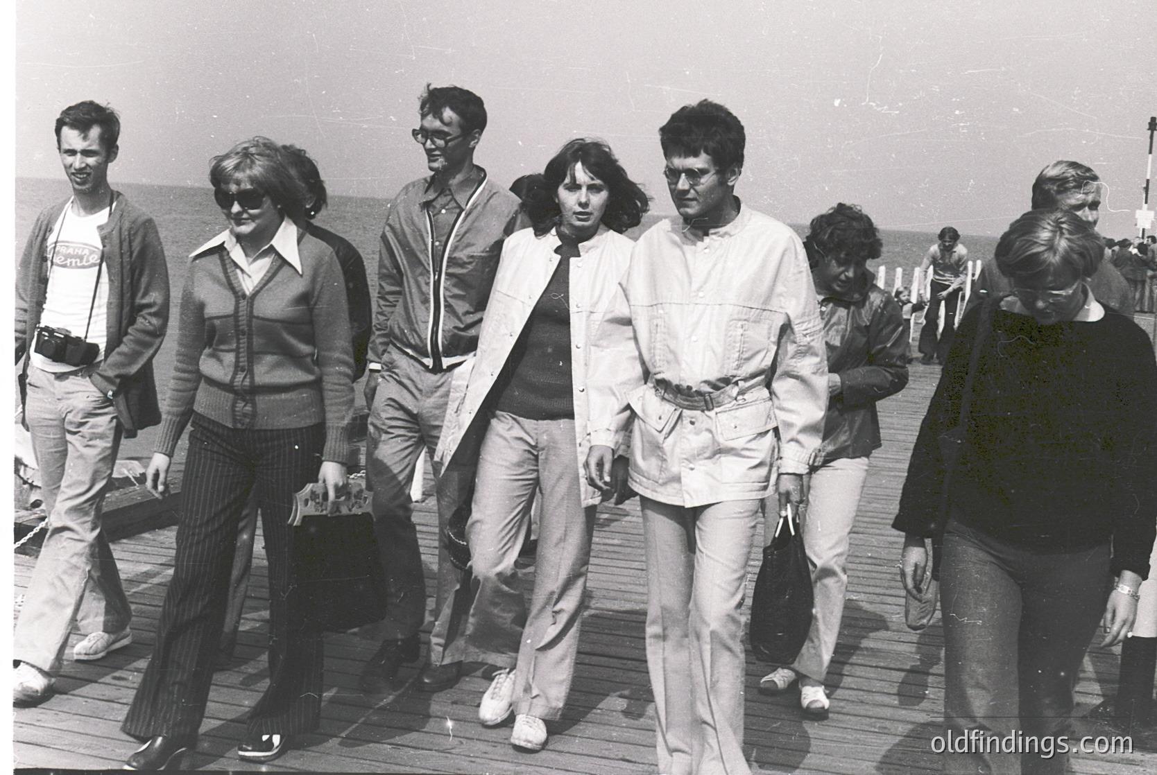 Group of seven individuals in 1970s beachwear strolling on a wooden pier, likely a seaside resort. Men wear wide-legged trousers, button-ups, and sunglasses; women in knee-length dresses or skirts with cardigans. Casual, relaxed summer attire suggests leisure activity.